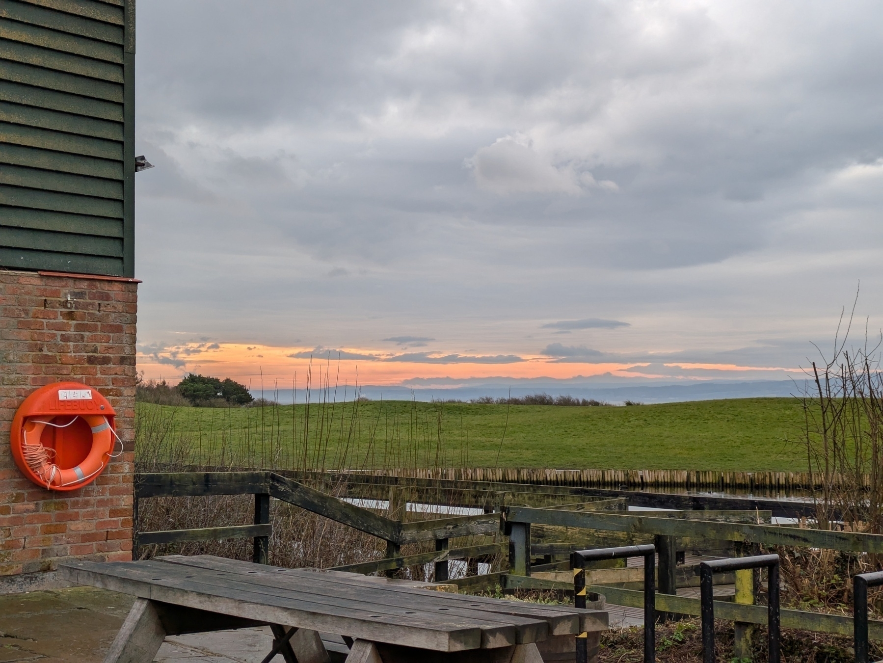 A wooden picnic table sits near a brick building with a lifebuoy, overlooking a grassy field and a cloudy rose-hued sky.