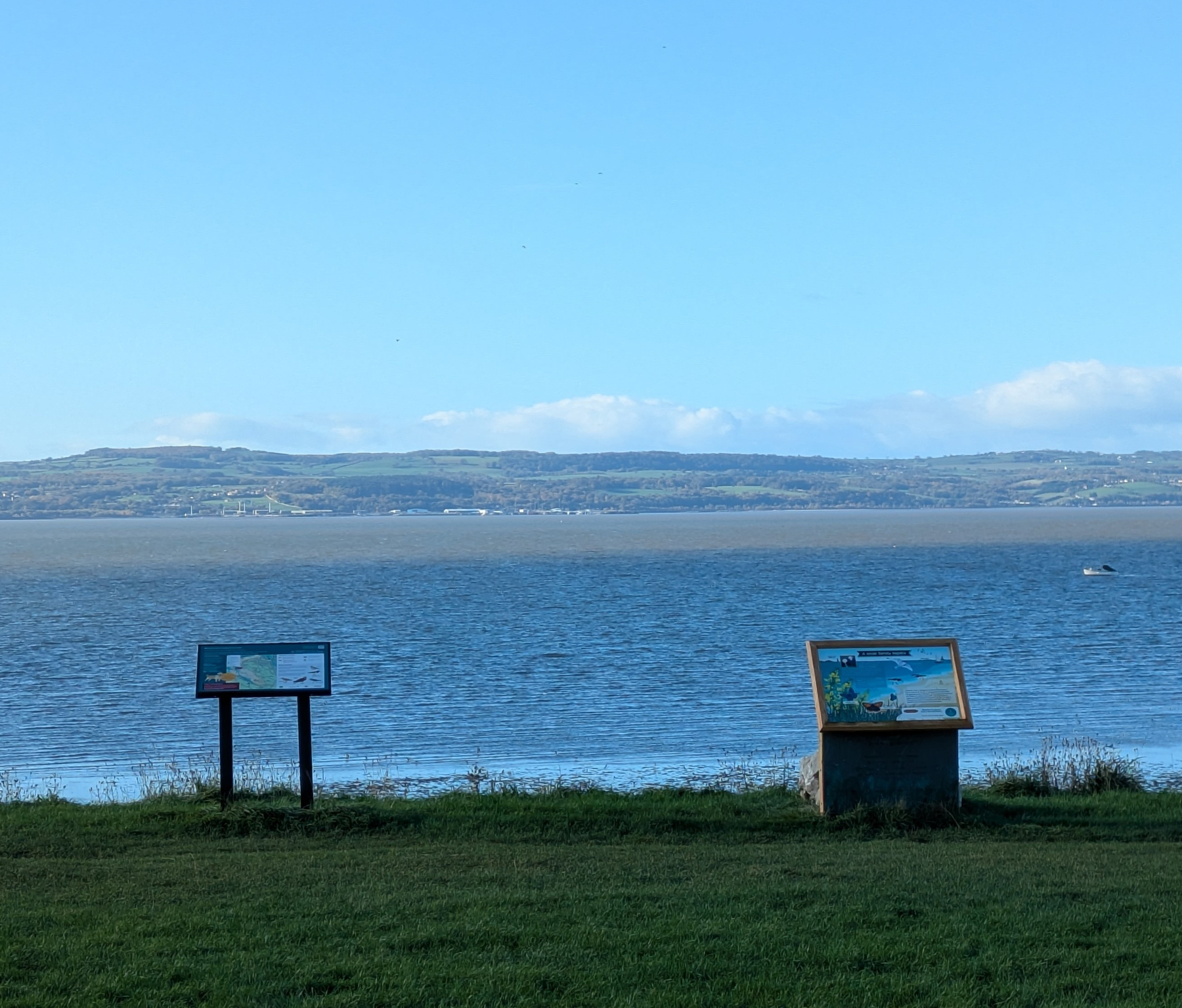 A peaceful coastal scene features grassy land in the foreground, informational signs, a calm body of water, and distant hills under a blue sky.
