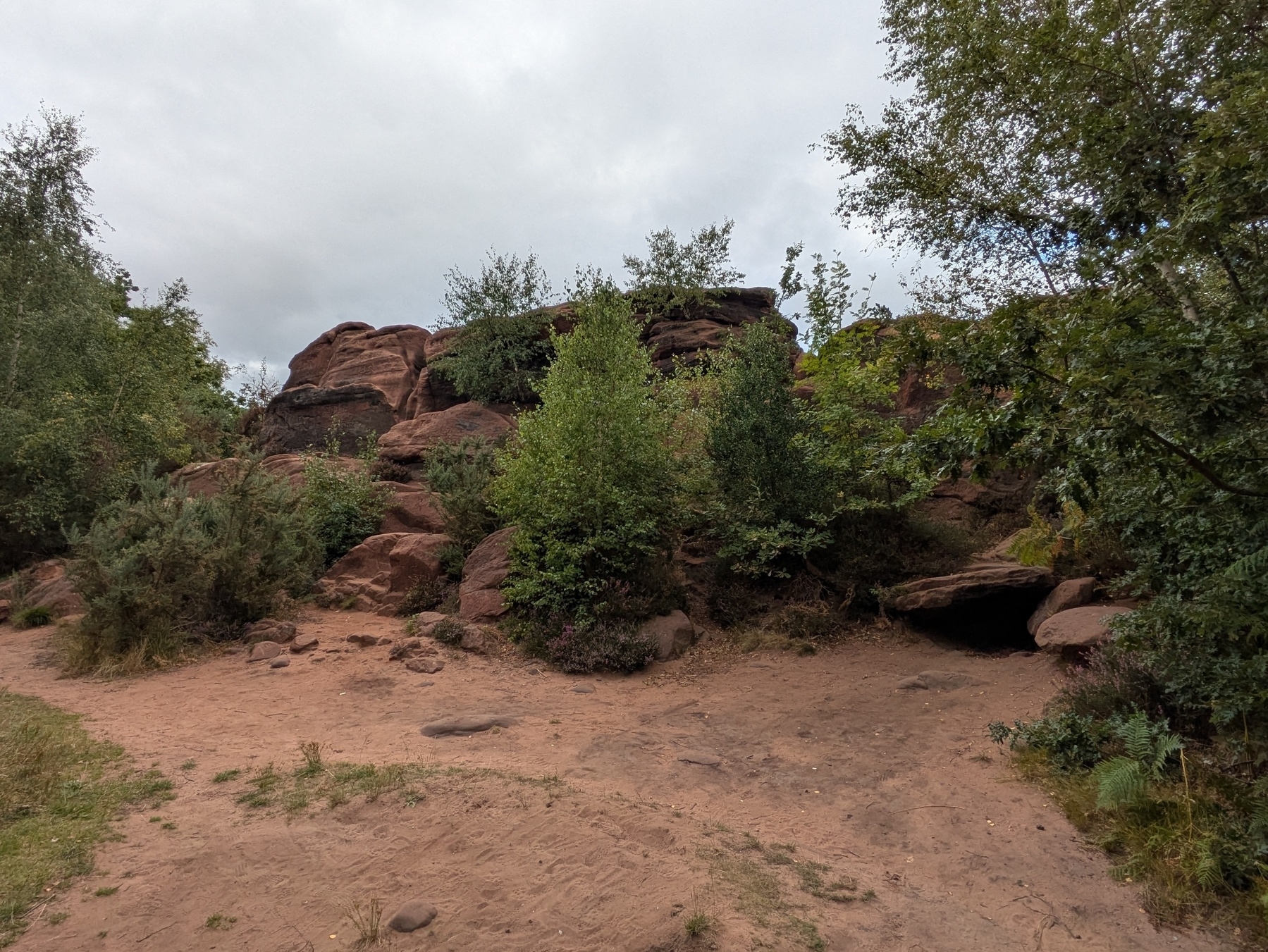 A sandy path leads to a rocky area surrounded by lush greenery and trees under a cloudy sky.