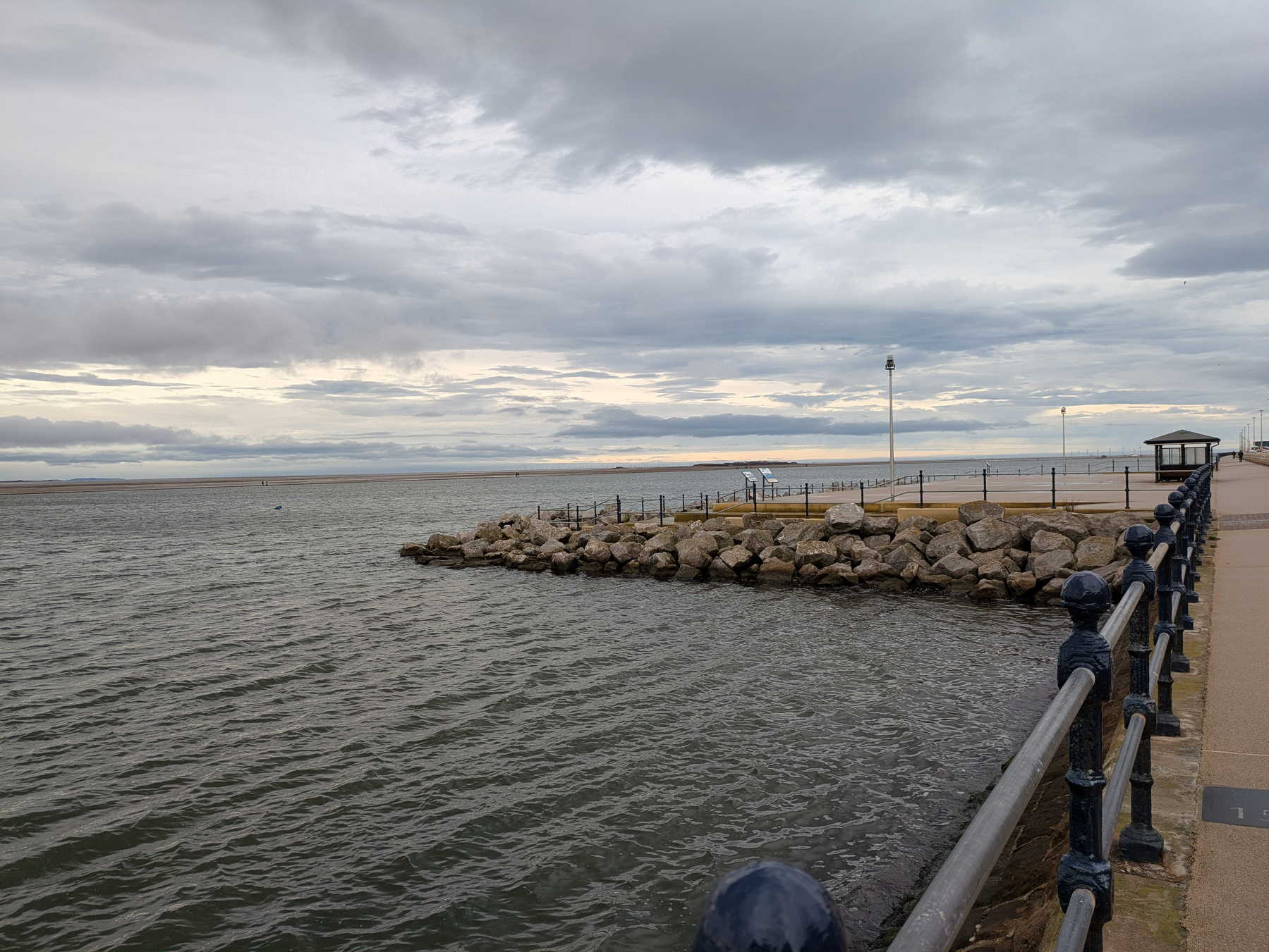 A rocky shoreline is bordered by a railing along a coastal walkway under a cloudy sky.