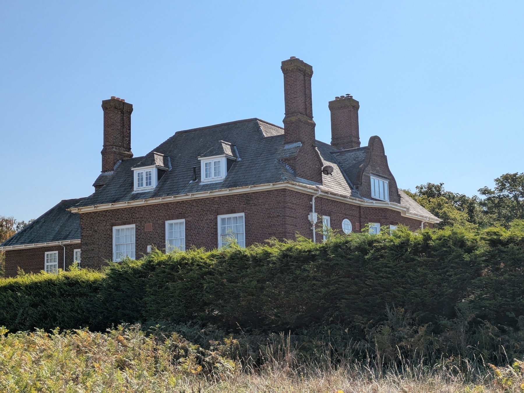 A large brick house with multiple chimneys is partially obscured by tall green hedges under a clear blue sky.