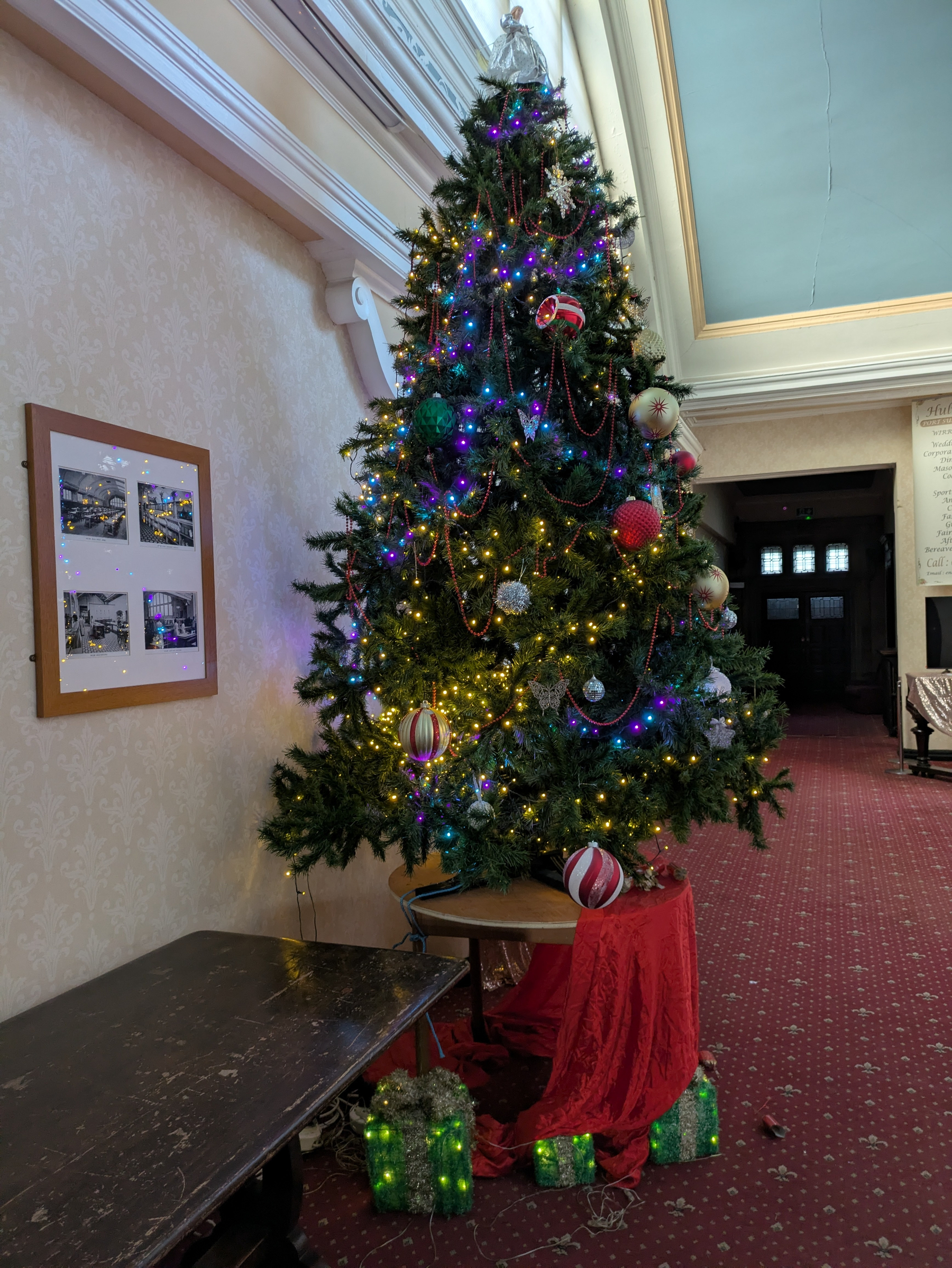 A decorated Christmas tree with lights, ornaments, and a red skirt is surrounded by wrapped presents in a carpeted room.