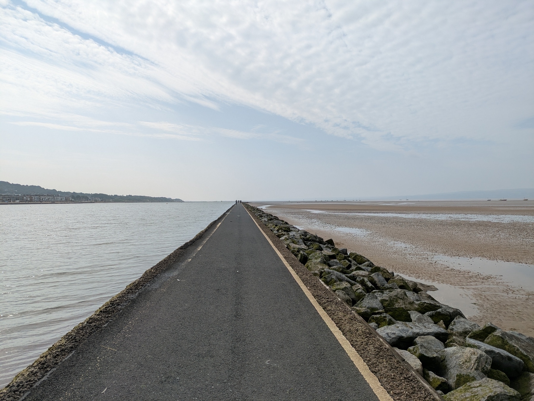 A long, narrow path extends between the sea and a rocky shoreline under a partly cloudy sky.