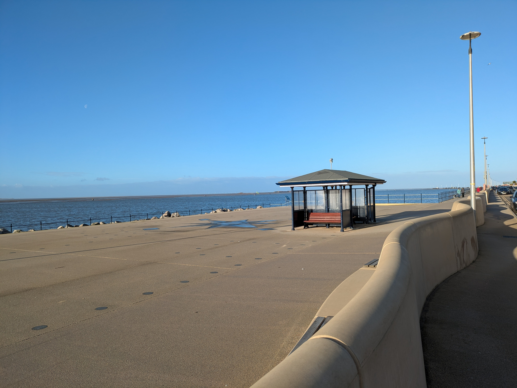 A seaside promenade features a walkway with a sheltered bench overlooking a calm sea under a clear blue sky.