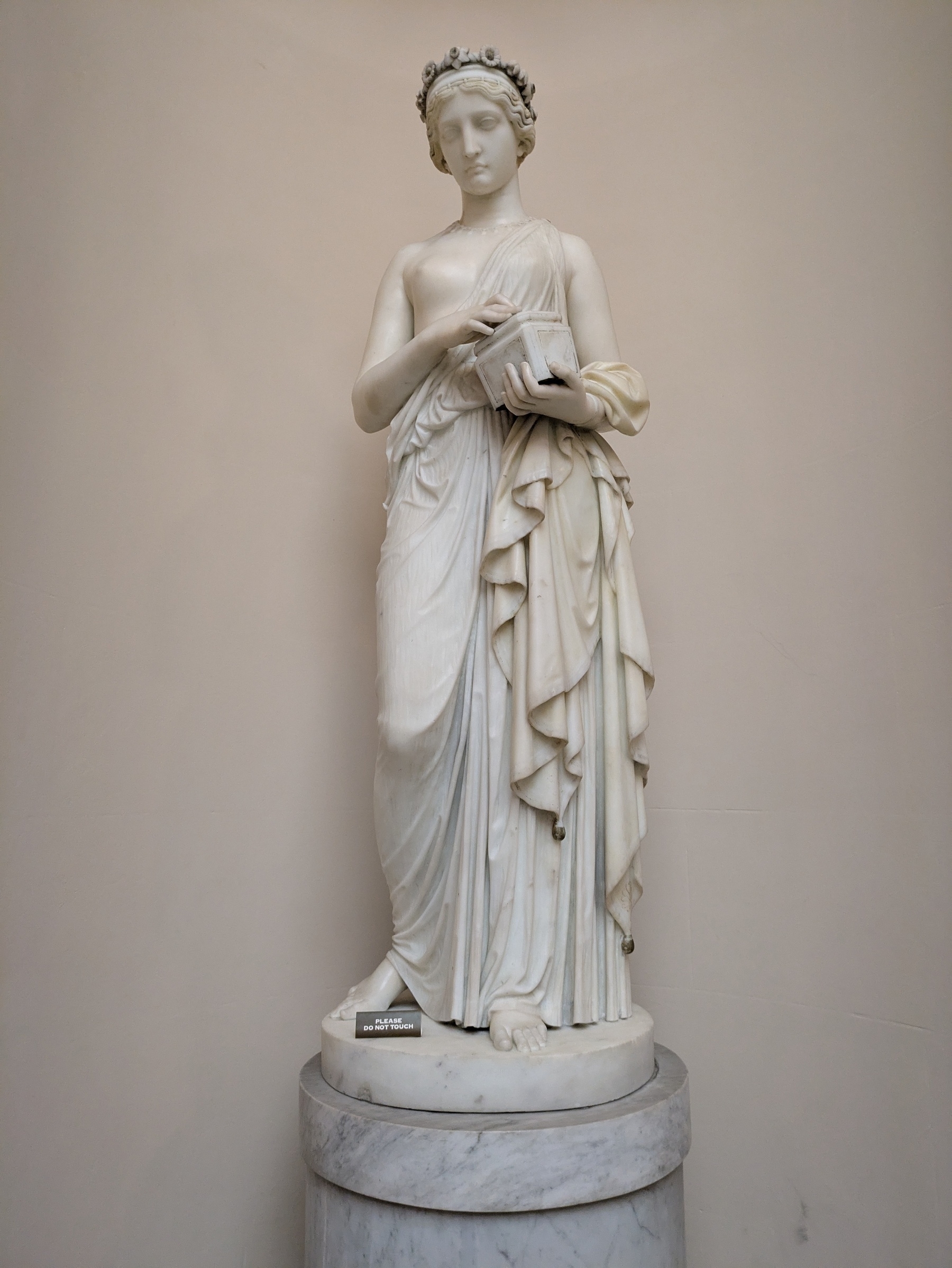 A classical marble statue depicts a draped figure holding an object, standing gracefully on a pedestal.