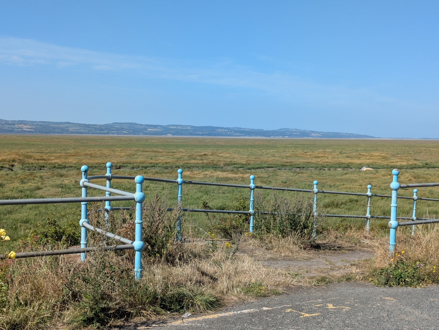 A coastal landscape features grassy fields stretching toward distant hills, bordered by a blue metal railing.