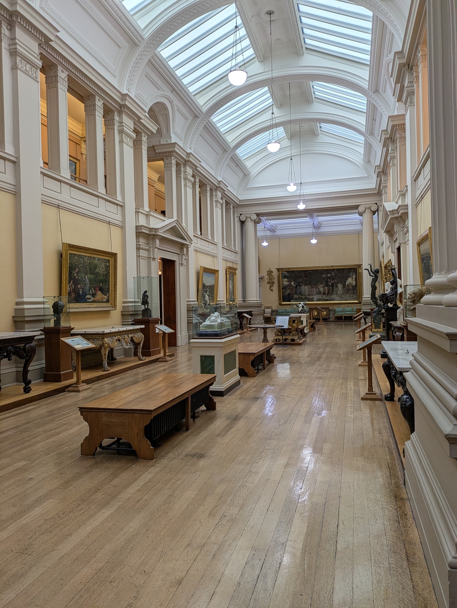 A spacious, well-lit gallery features wooden benches, sculptures, and classical paintings lining the walls.