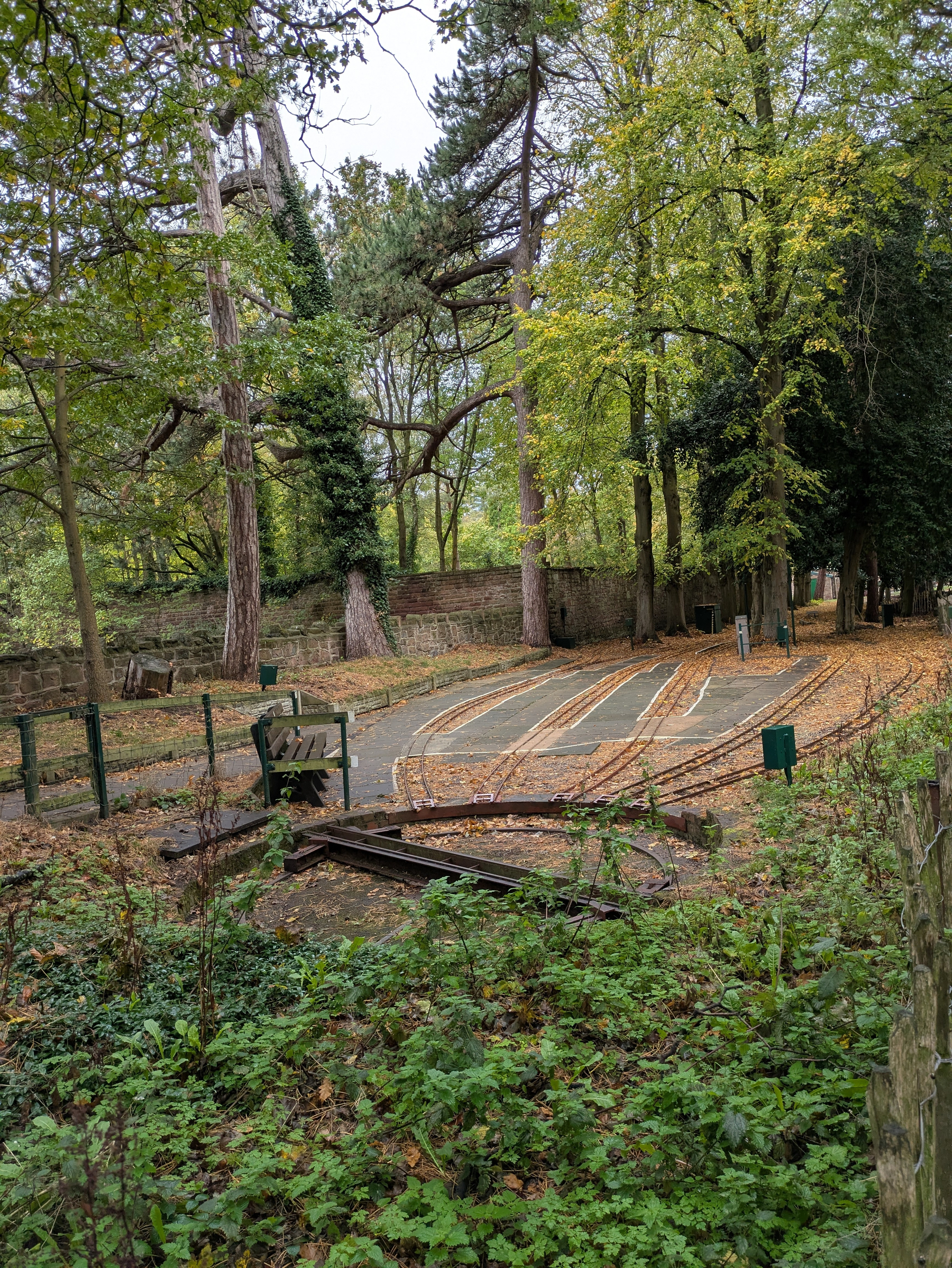 A serene forest scene features a turntable leading to a station for a miniature railway whose tracks are surrounded by scattered fallen leaves.