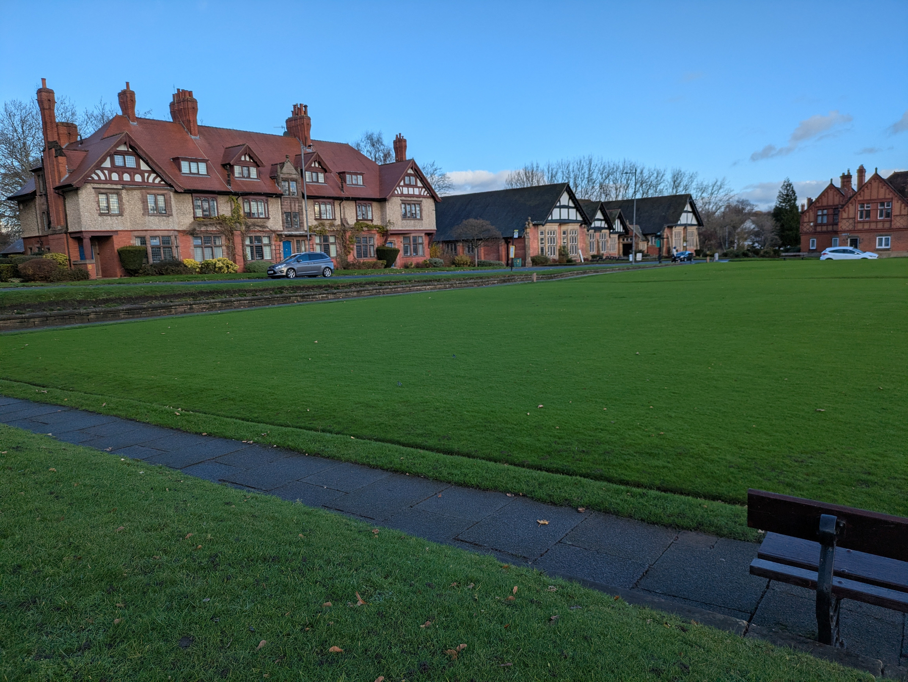 A grassy lawn is bordered by a path, with a row of traditional houses in the background and a bench in the foreground.