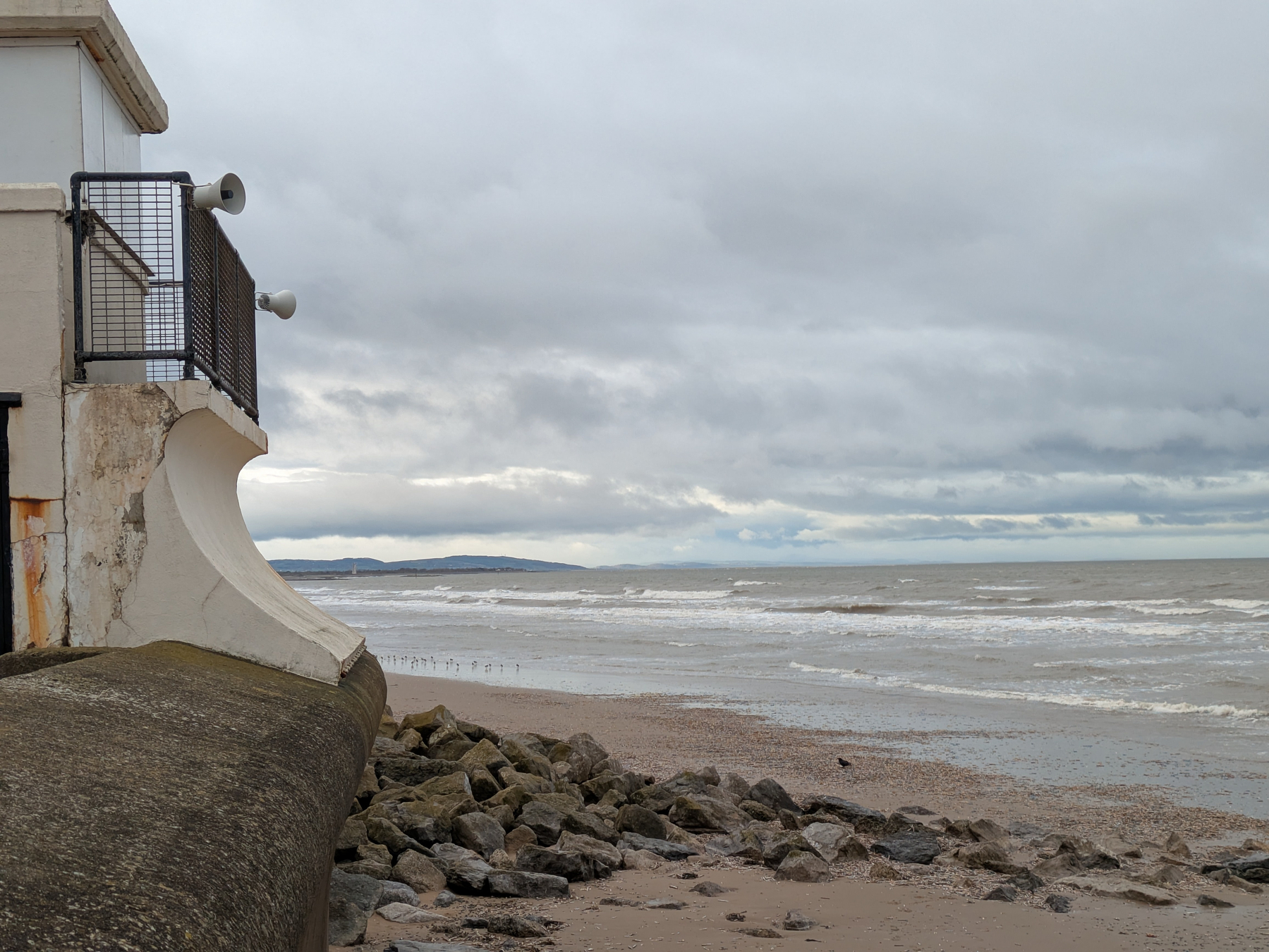 A rocky beach with a concrete structure overlooks a cloudy and wavy coastline.