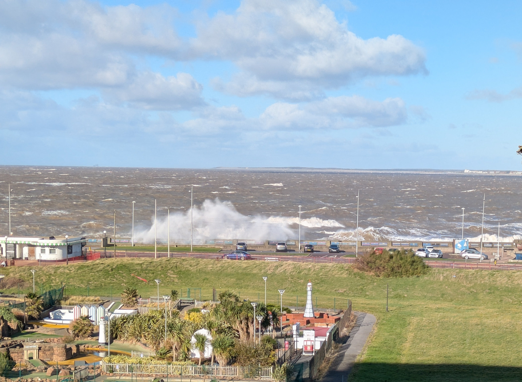 Waves crash against the shoreline while cars drive along a coastal road under a partly cloudy sky.