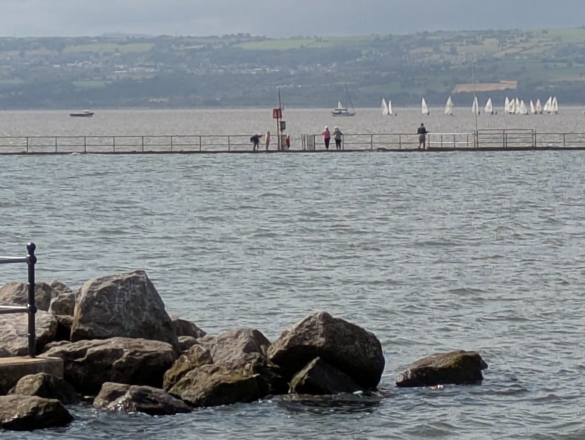 Marine lake with rocks in the foreground, people walking around the lake in the middle distance, then yachts in the estuary and Welsh hills beyond.