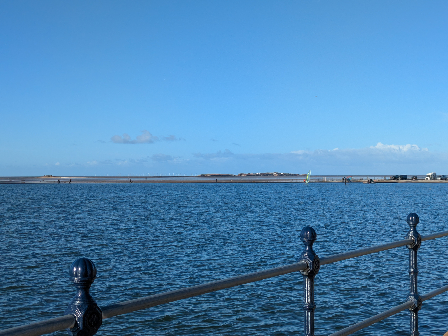 A calm waterfront scene features a railing in the foreground and a distant shoreline under a clear blue sky.