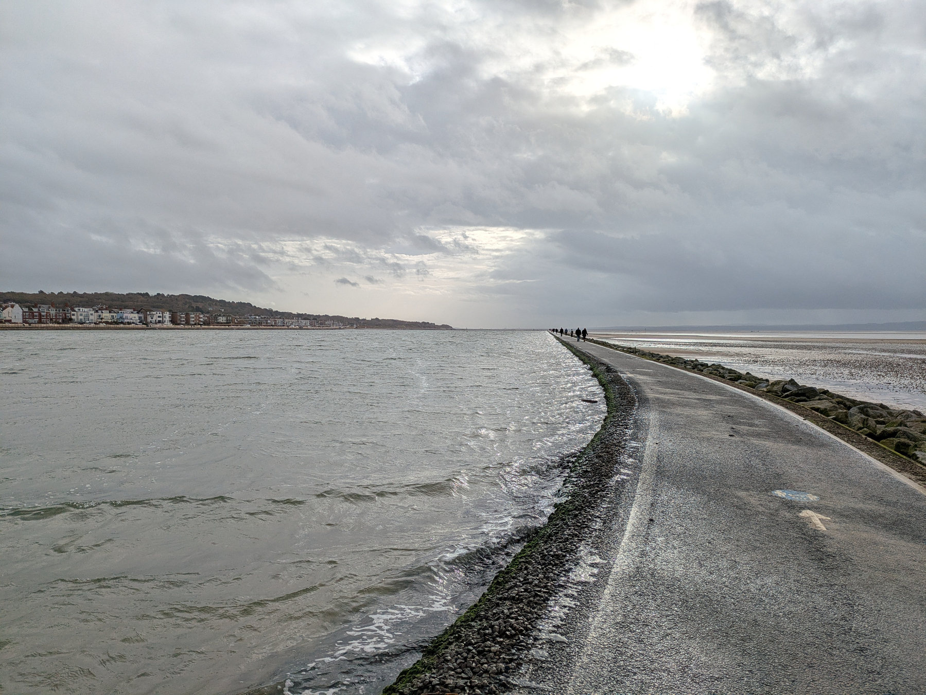 A narrow road flanked by water leads towards the horizon under a cloudy sky.