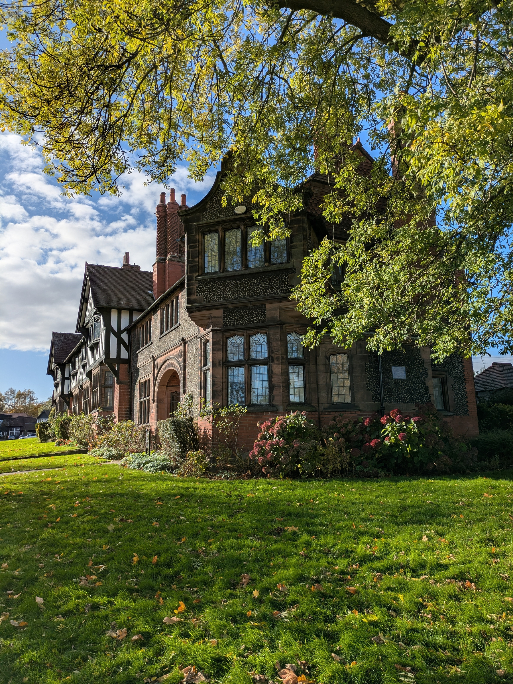 A large, historic brick house with intricate designs is surrounded by lush greenery and under a clear blue sky.