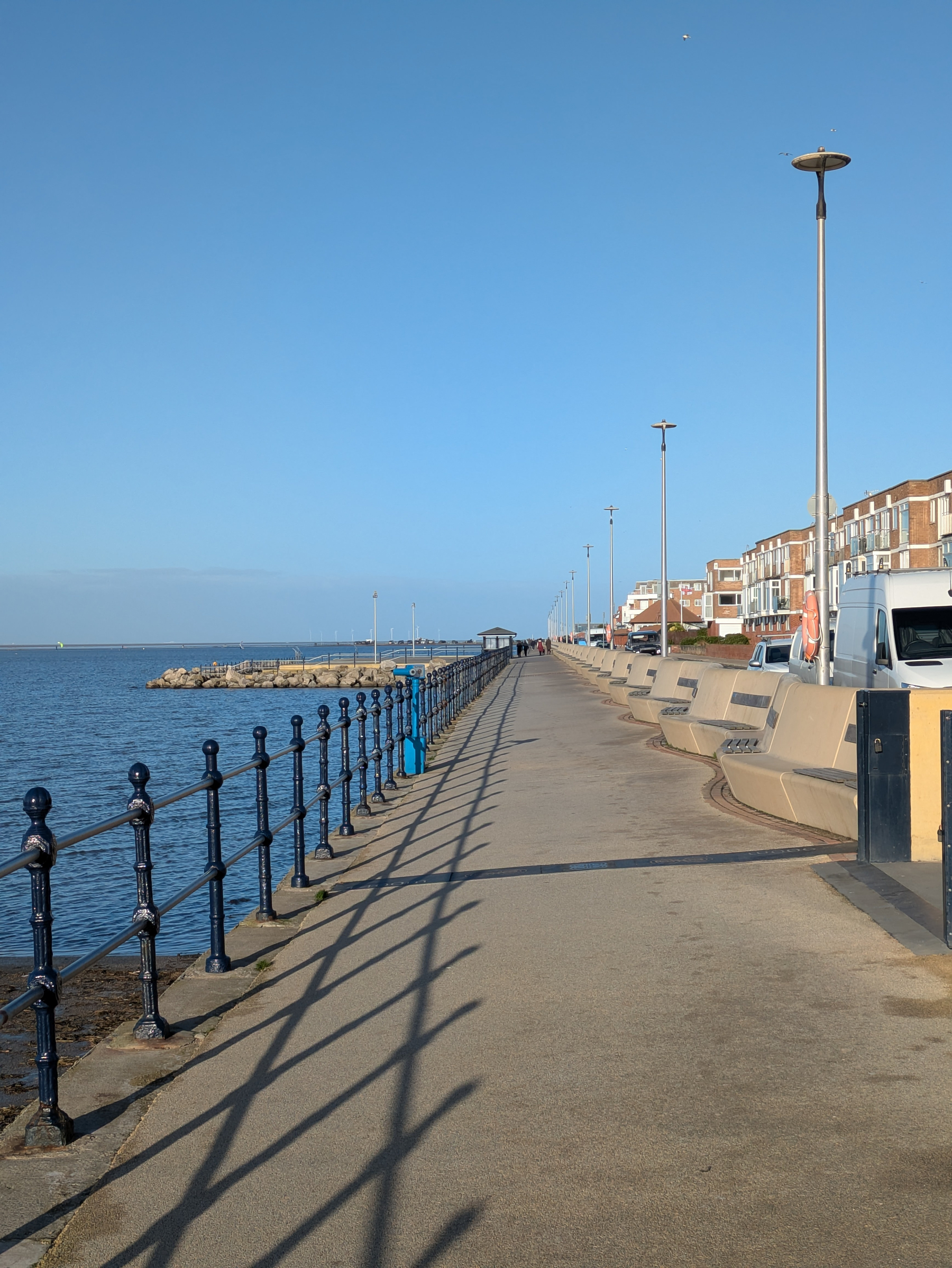 A seaside promenade with a long path, metal railings, streetlights, and buildings on the right under a clear blue sky.