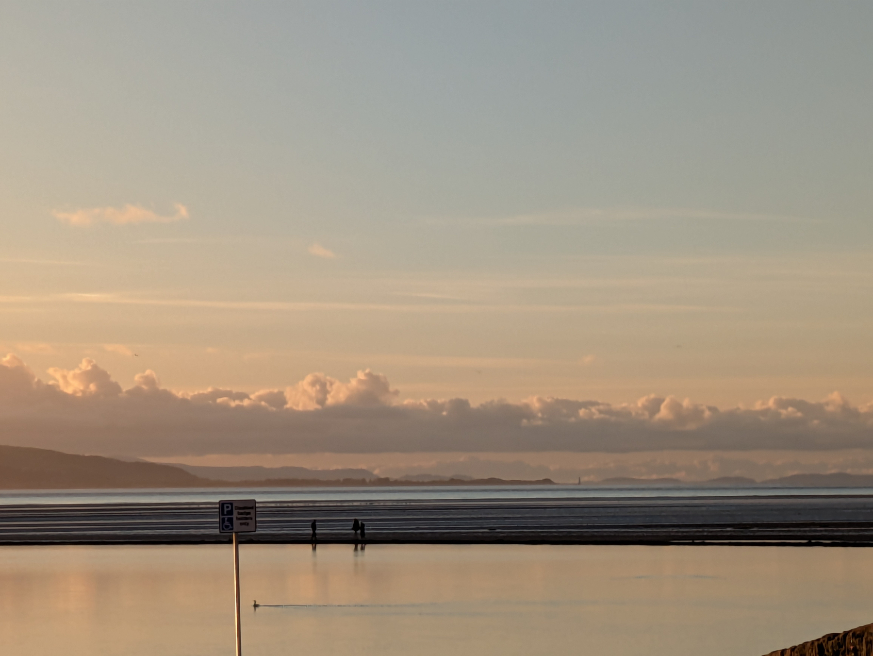 A tranquil scene features a vast body of water reflecting the golden hues of the sunset, with distant hills and fluffy clouds under a clear sky.