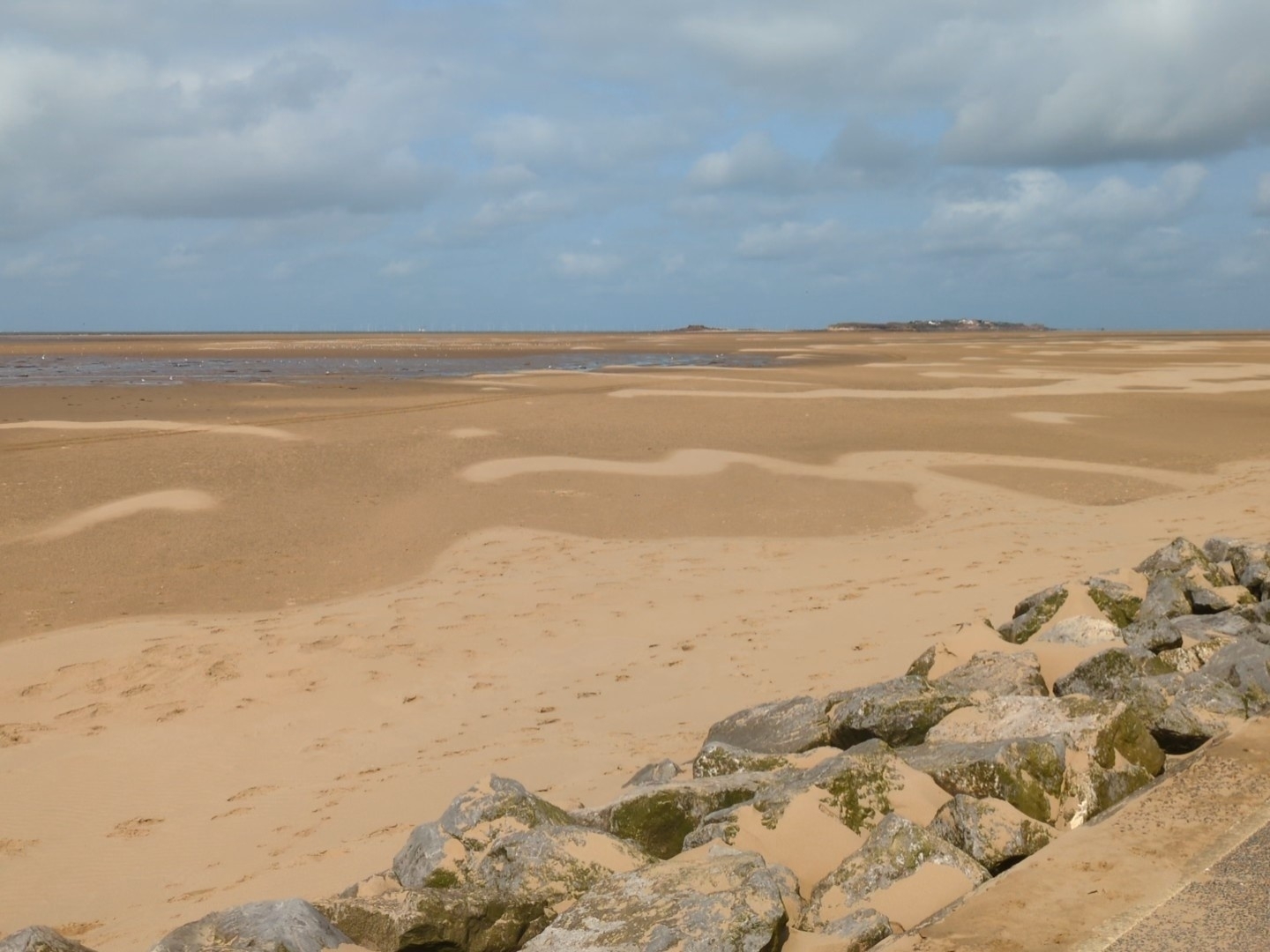 A vast sandy beach stretches into the distance under a partly cloudy sky, with large rocks in the foreground.