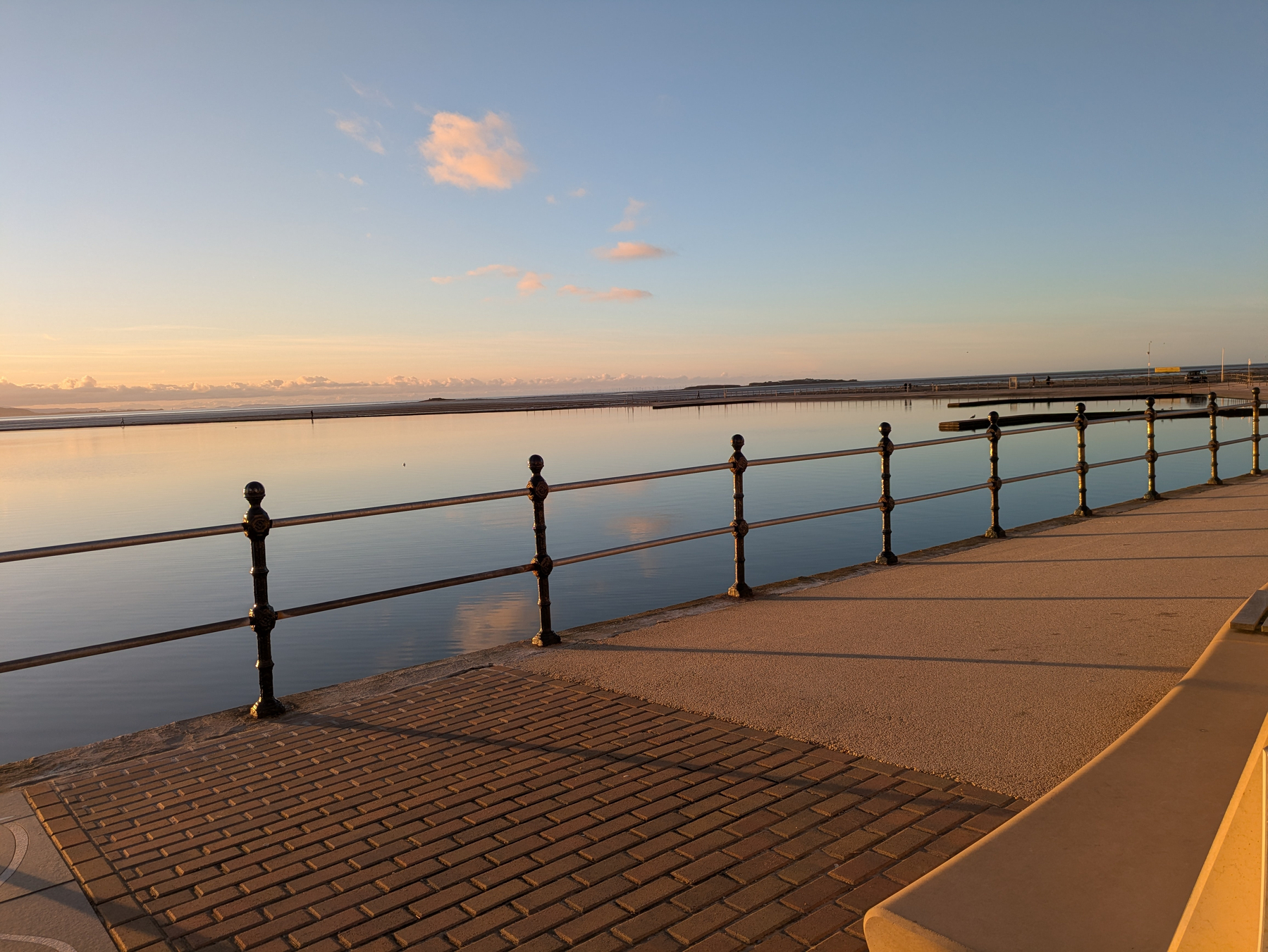 A peaceful waterfront scene at sunset features a calm body of water, a metal railing, and a paved walkway.