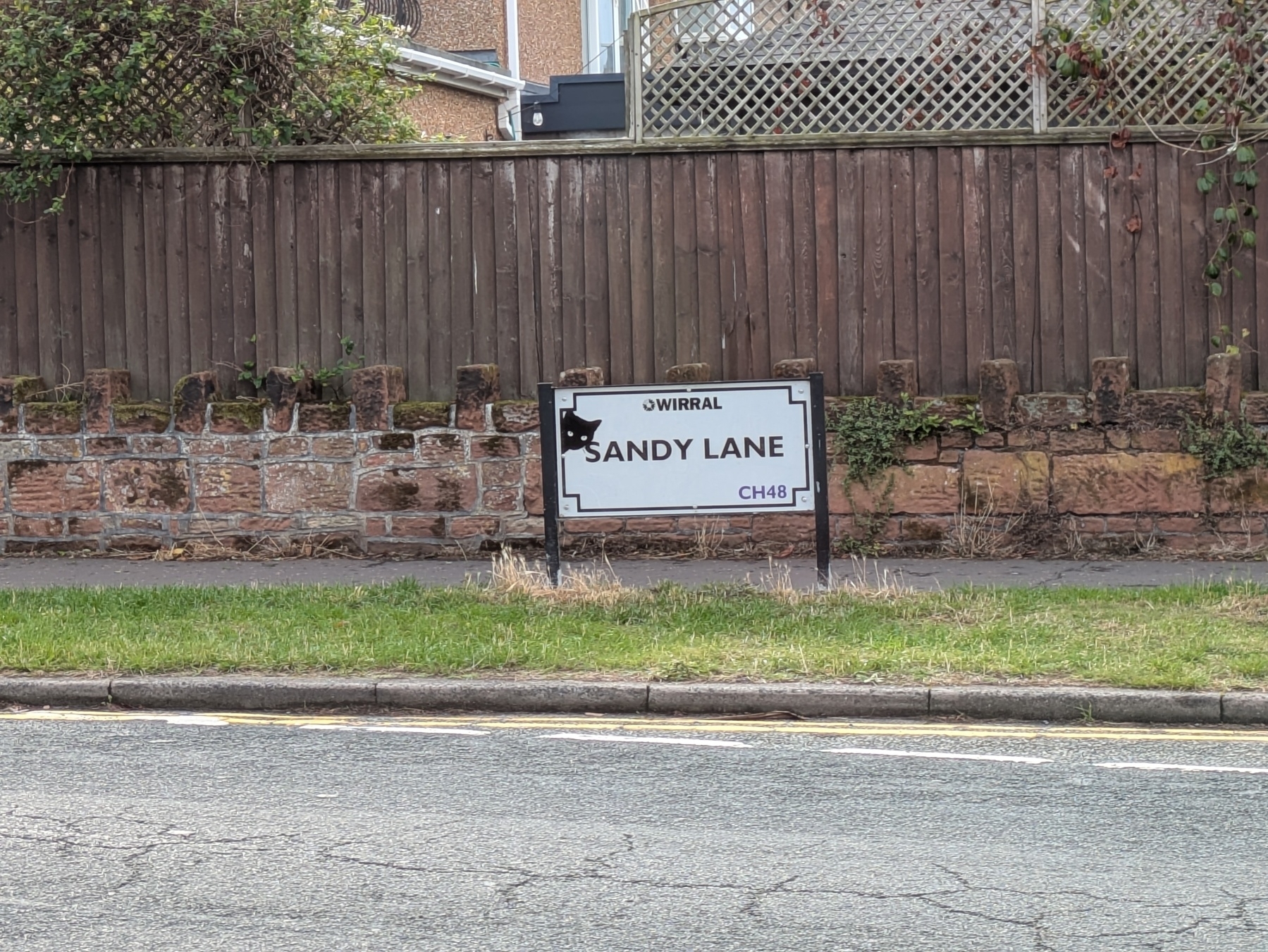 A street sign for Sandy Lane in Wirral is displayed in front of a wooden and brick fence.