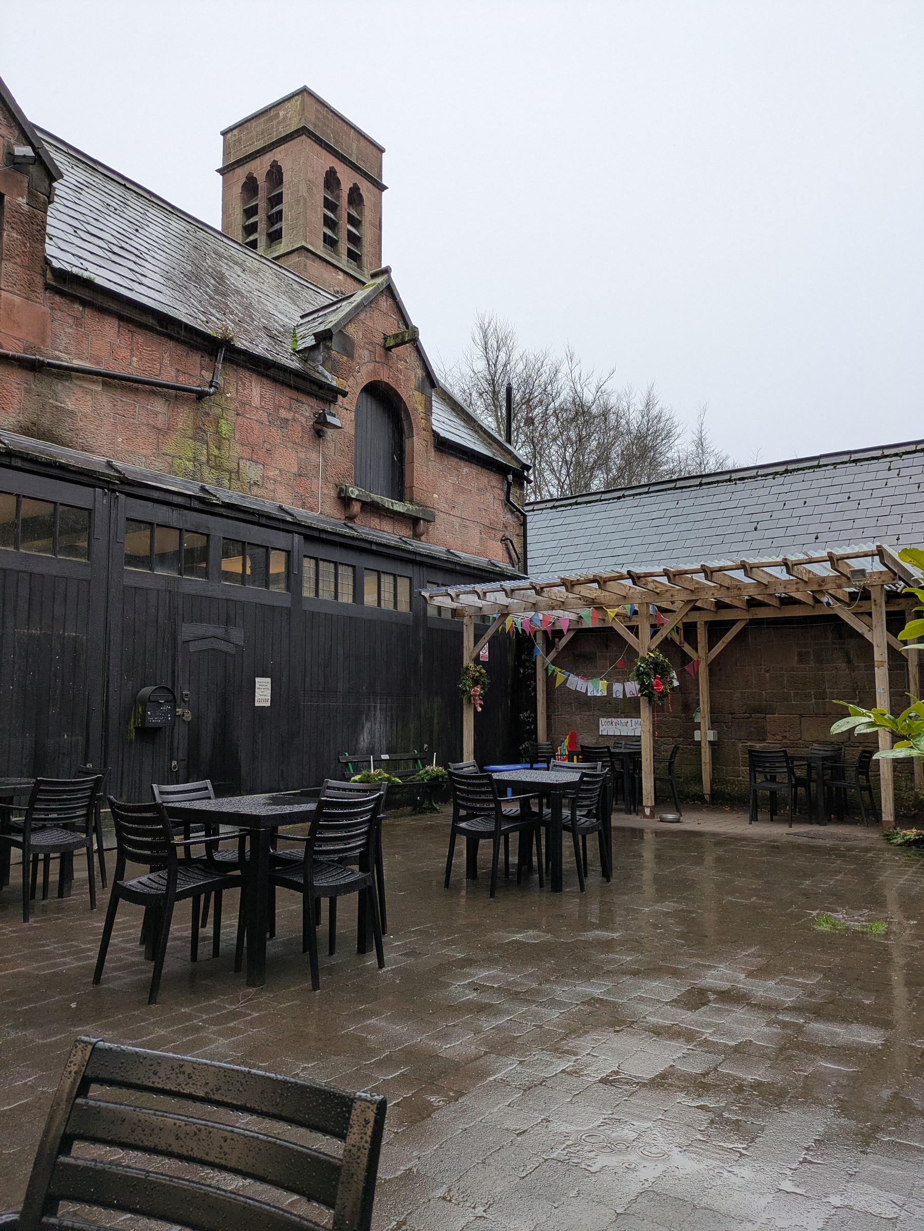 A quaint outdoor courtyard with tables and chairs sits wet under overcast skies, adjacent to a rustic brick building with a tower.