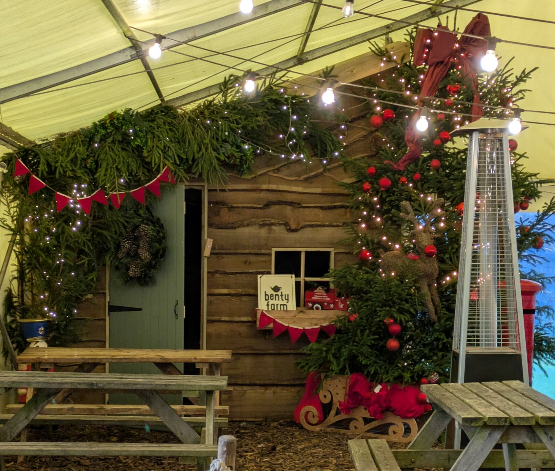 A festive, rustic scene features a wooden cabin adorned with Christmas decorations, including a tree with ornaments, garlands, and string lights, under a lit tent with picnic tables and a heater.