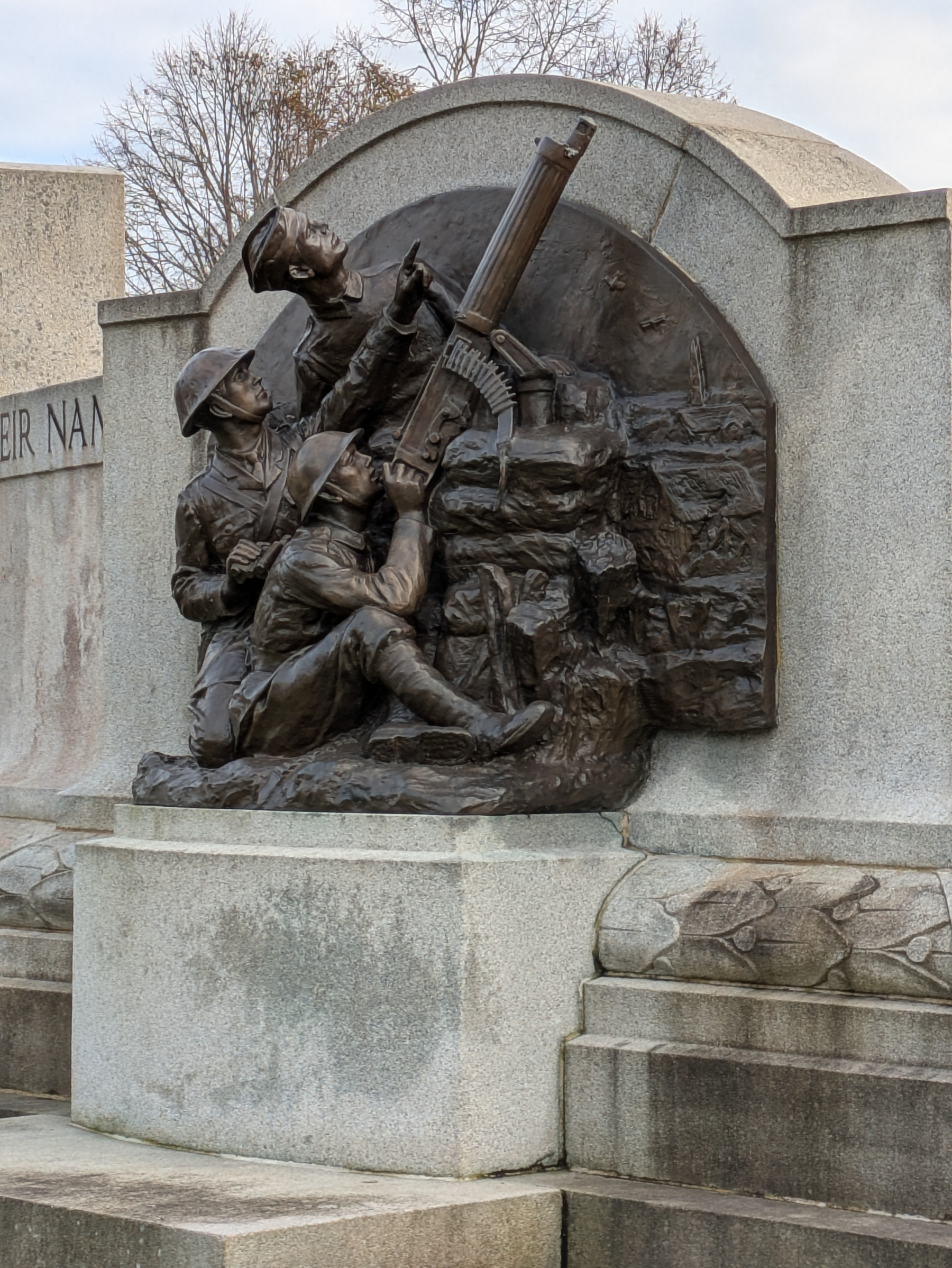 A bronze relief sculpture depicts two soldiers operating a machine gun atop a stone monument.