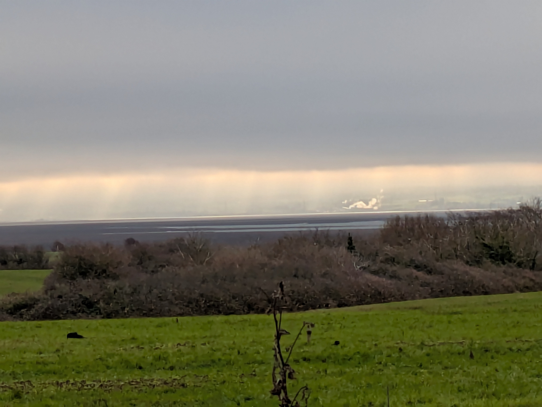 A scenic landscape features green fields, distant trees, a body of water, and a cloudy sky with sun rays peeking through.