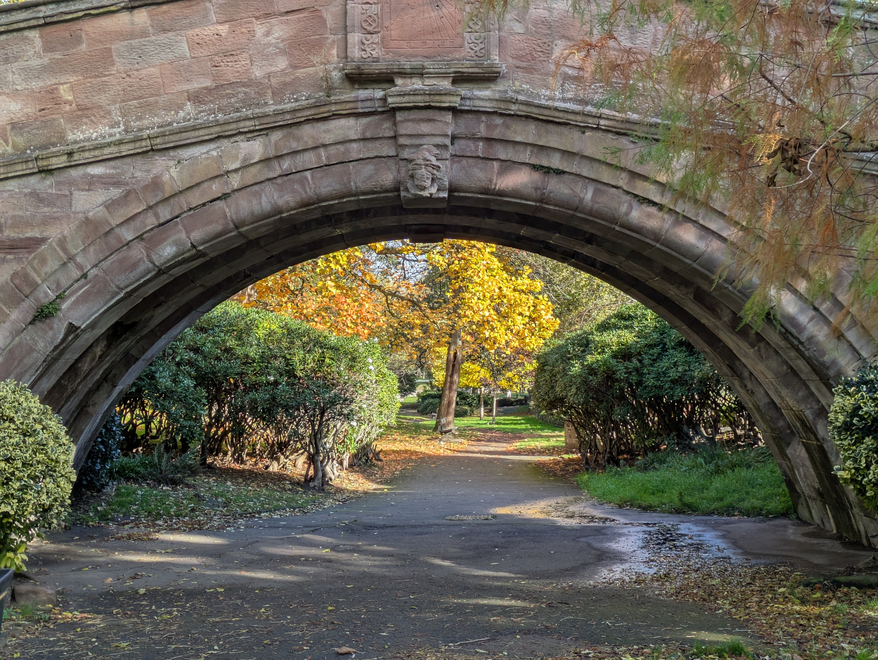 An arched stone bridge frames a path surrounded by lush greenery and trees with autumn foliage.