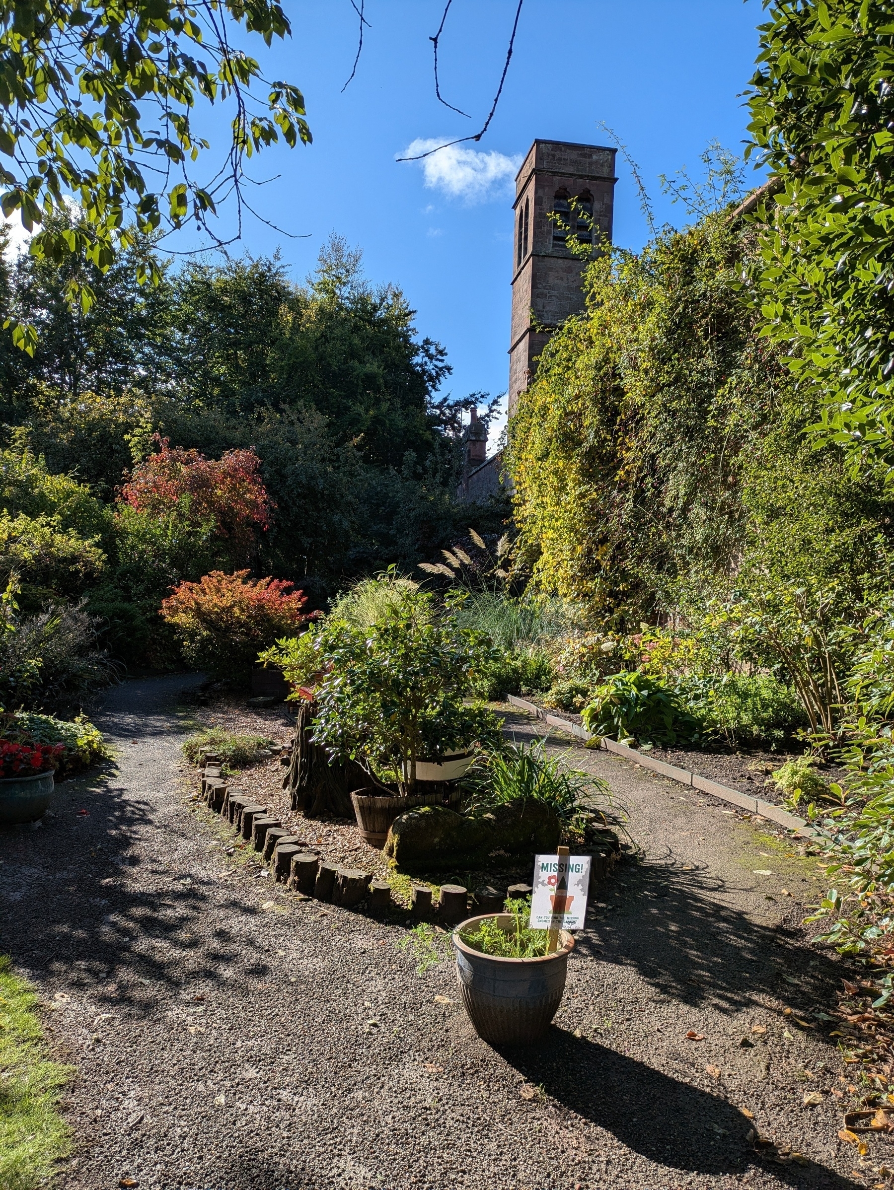 Edge of walled garden on right overlooked by tall stone tower. Wall covered with foliage. Pot with sign in foreground, shrubs and trees beyond, nearest ones in pots 