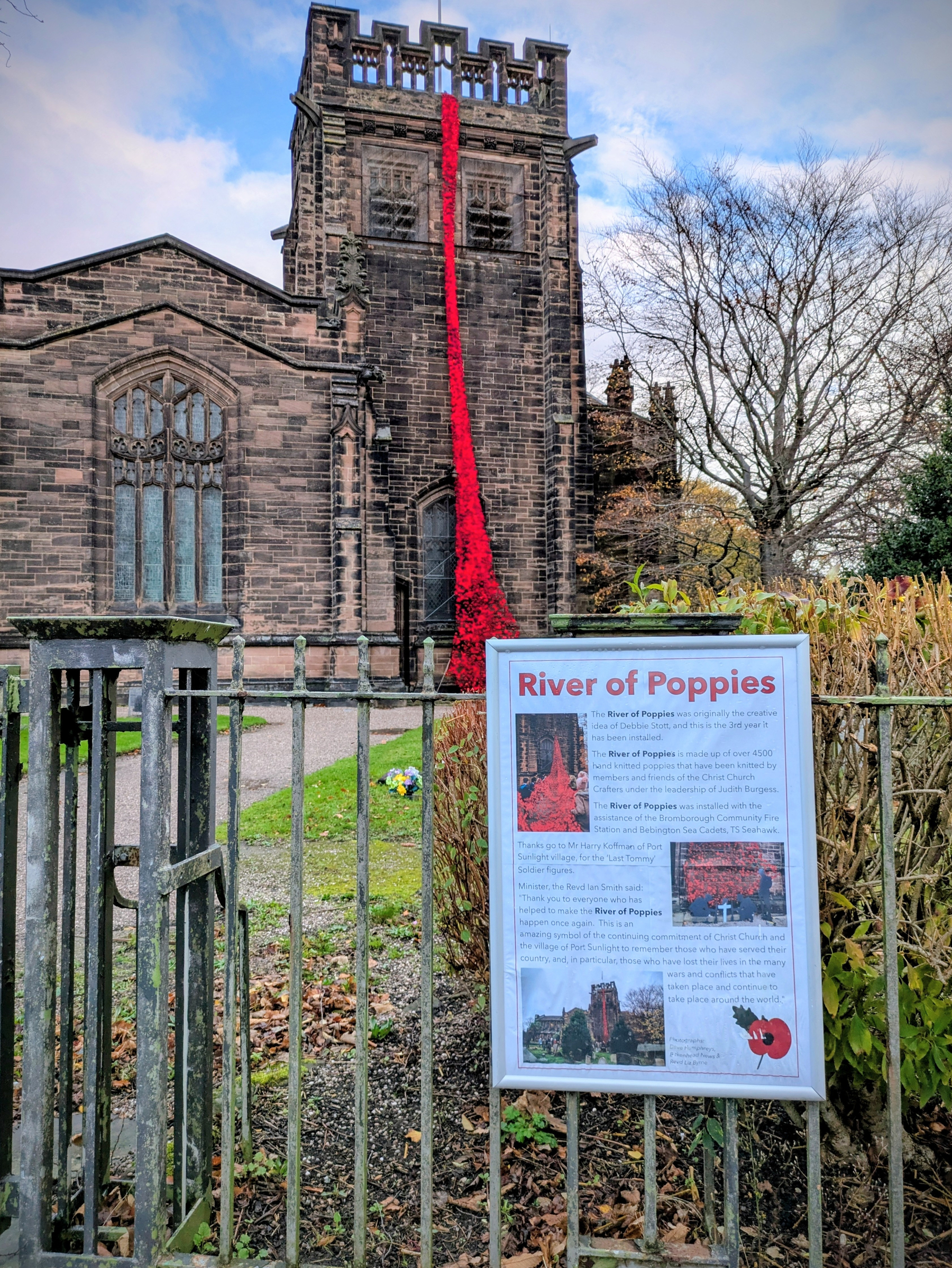 A historic stone church features a striking installation of cascading red poppies, with an informational sign titled River of Poppies in the foreground.