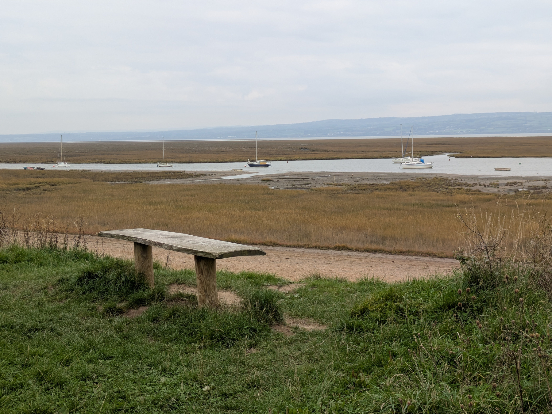 A rustic bench overlooks a tranquil marshland with several sailboats anchored in calm waters.