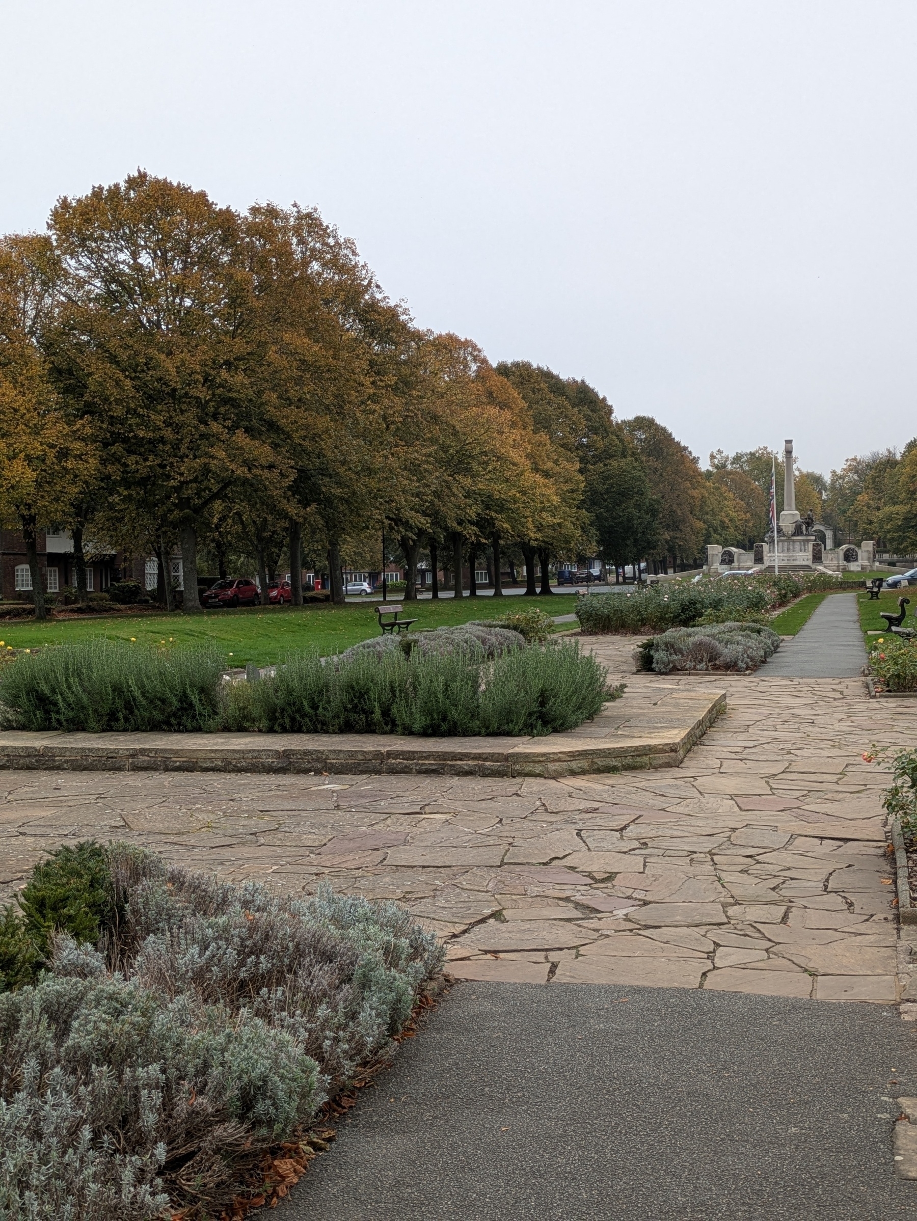 A paved walkway bordered by shrubs and trees leads to a distant monument in a park setting.