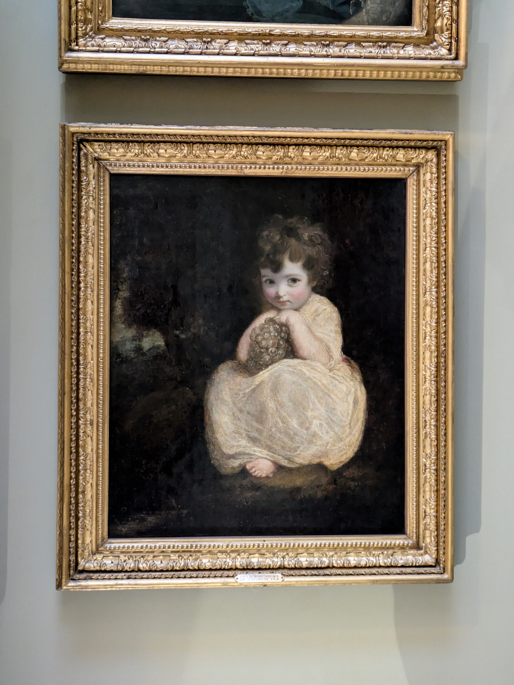 A young child in a light-colored dress sits holding a bunch of grapes in an ornate-framed painting.