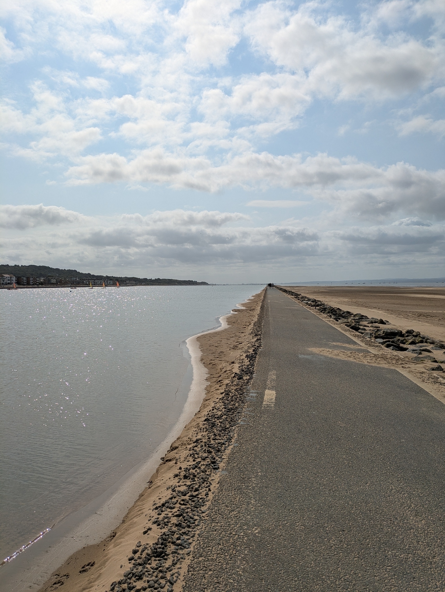 A narrow path flanked by a calm body of water on one side and a sandy beach on the other extends into the distance under a partly cloudy sky.