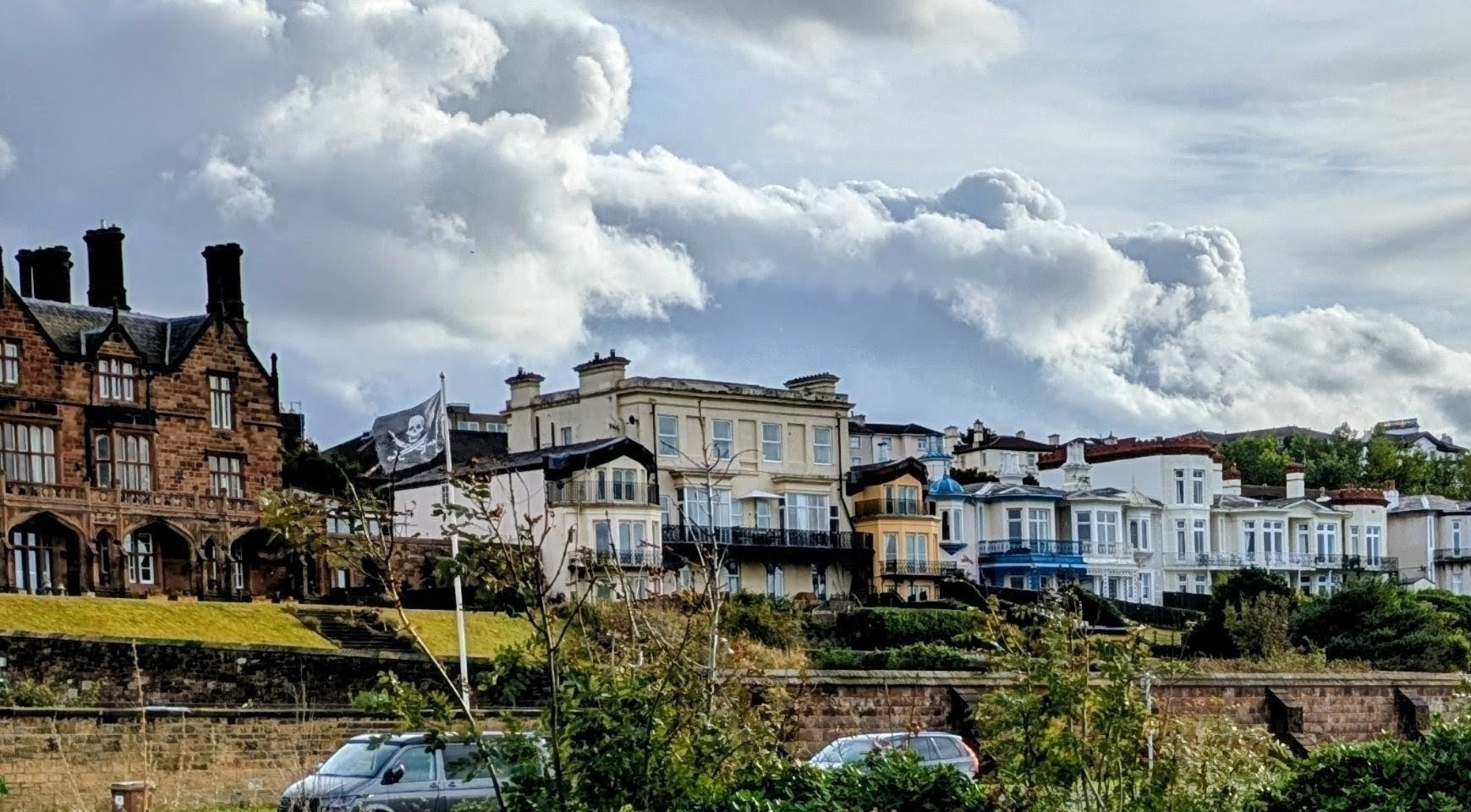 A row of historic buildings with ornate architecture is set against a backdrop of dramatic clouds, with parked cars in the foreground.