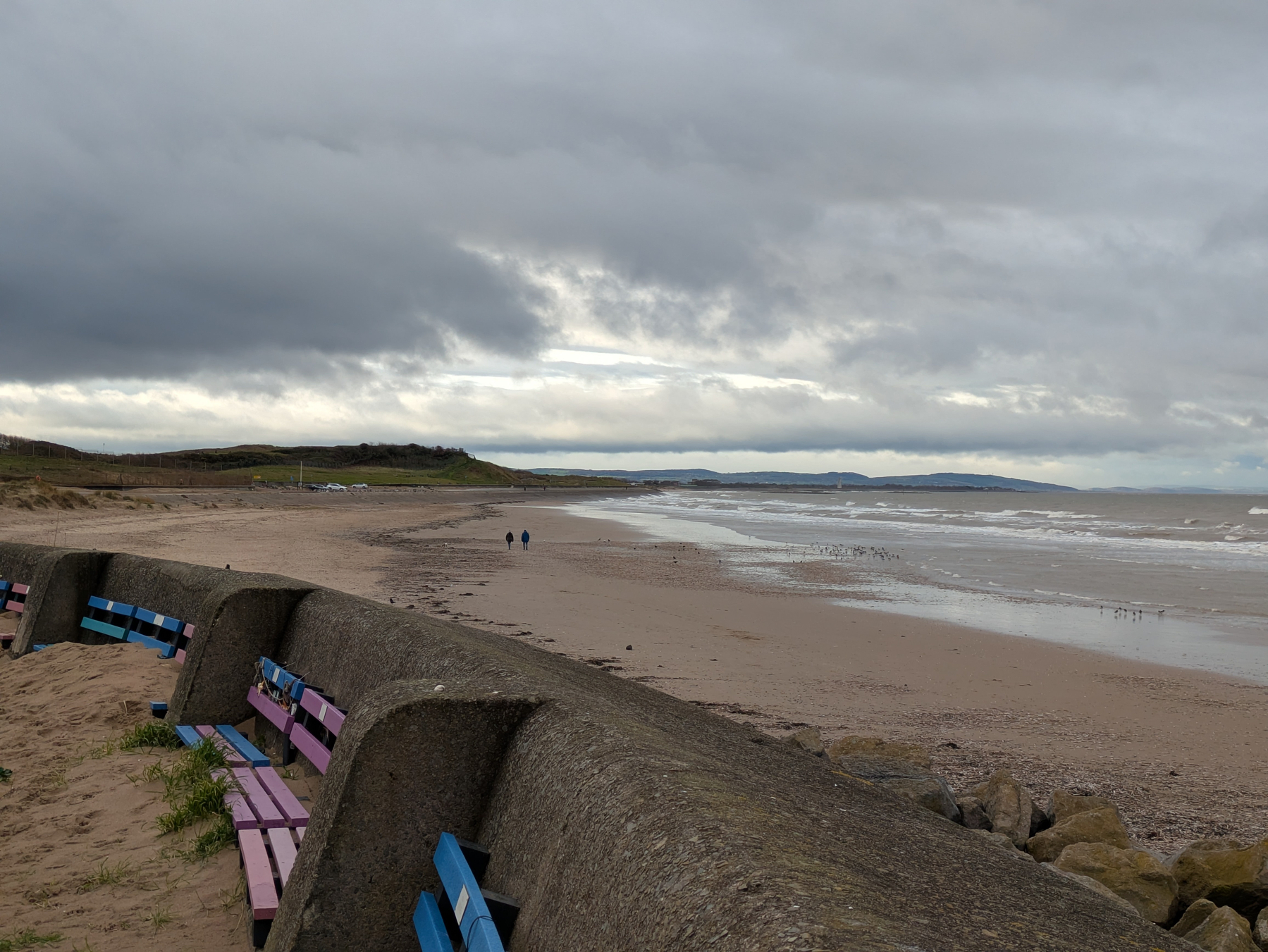 A coastal scene features a sandy beach, a curved sea wall, colorful benches, and two people walking under a cloudy sky.