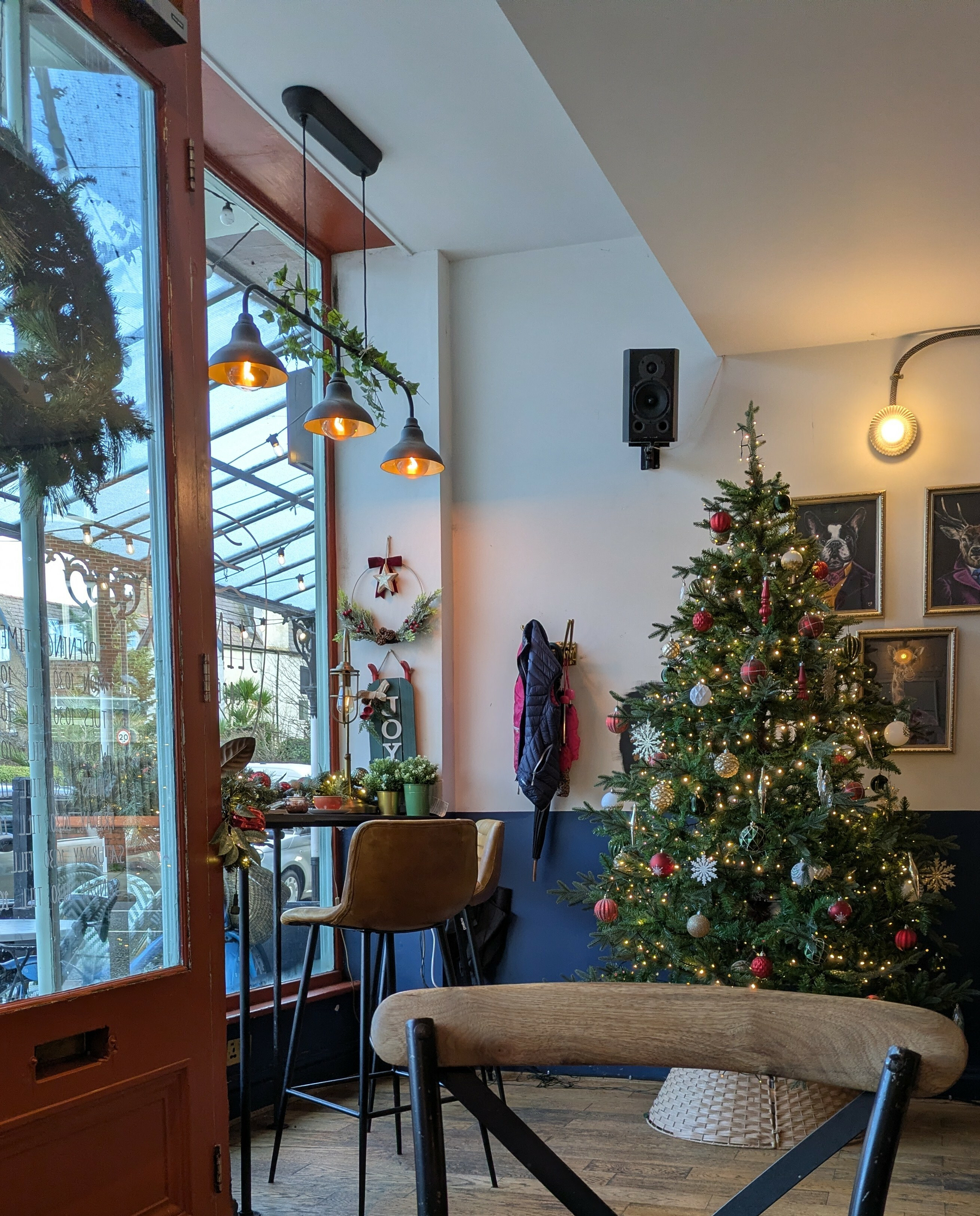 A cozy cafe features a decorated Christmas tree, a wreath on the door, and warm lighting creating a festive atmosphere.