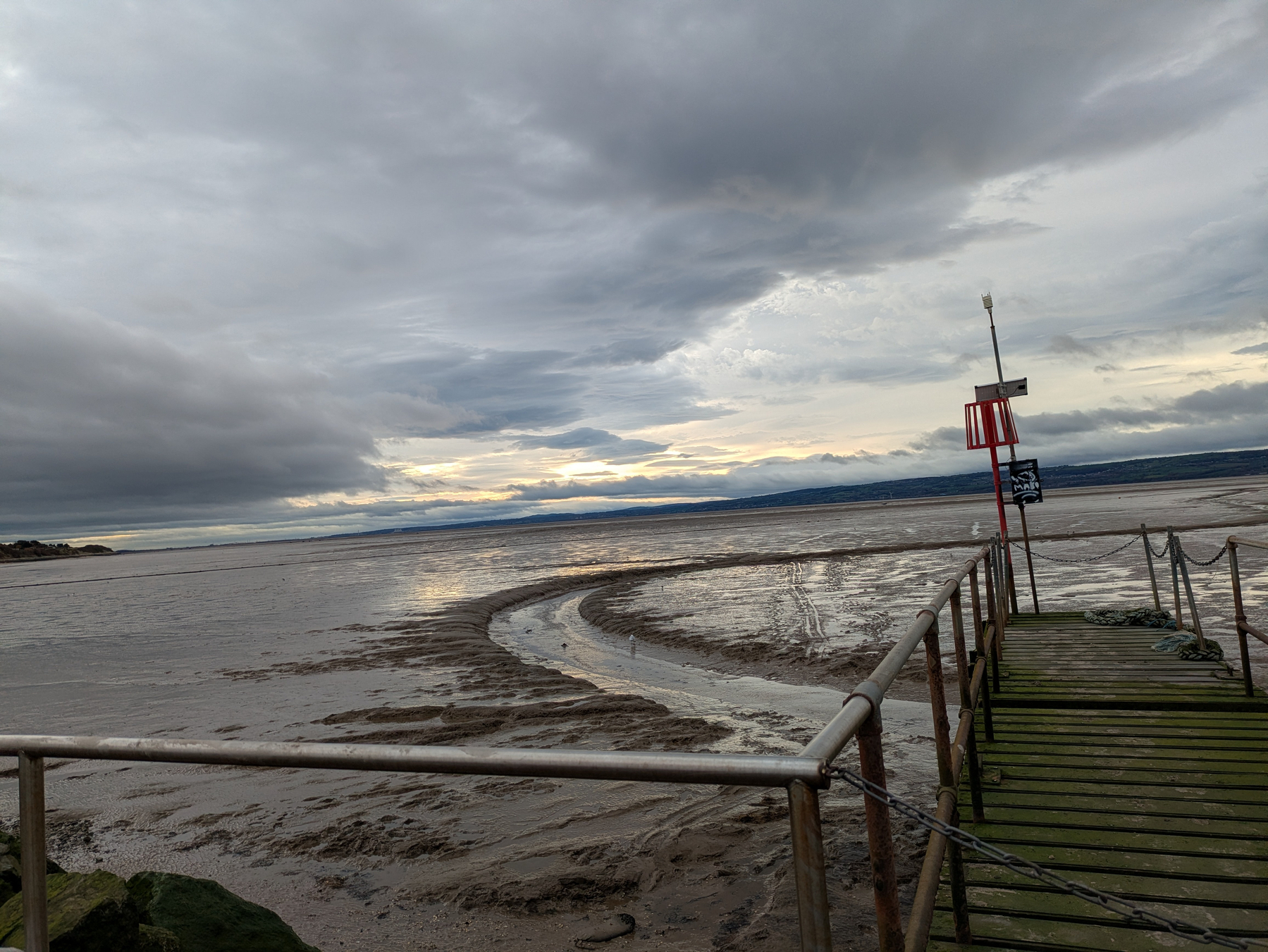 A wooden pier extends over a wide, muddy shoreline with a cloudy sky overhead.
