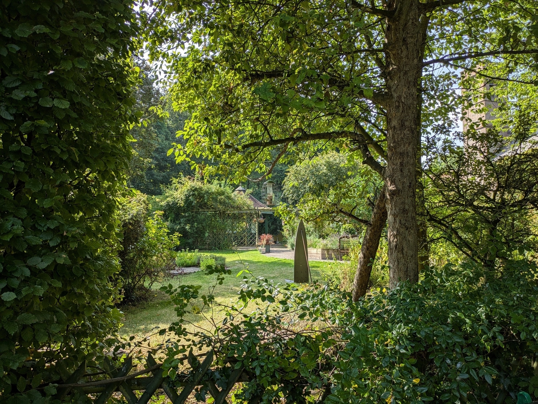 A lush garden features dense foliage, a pathway, and a distant structure surrounded by sunlight filtering through the trees.
