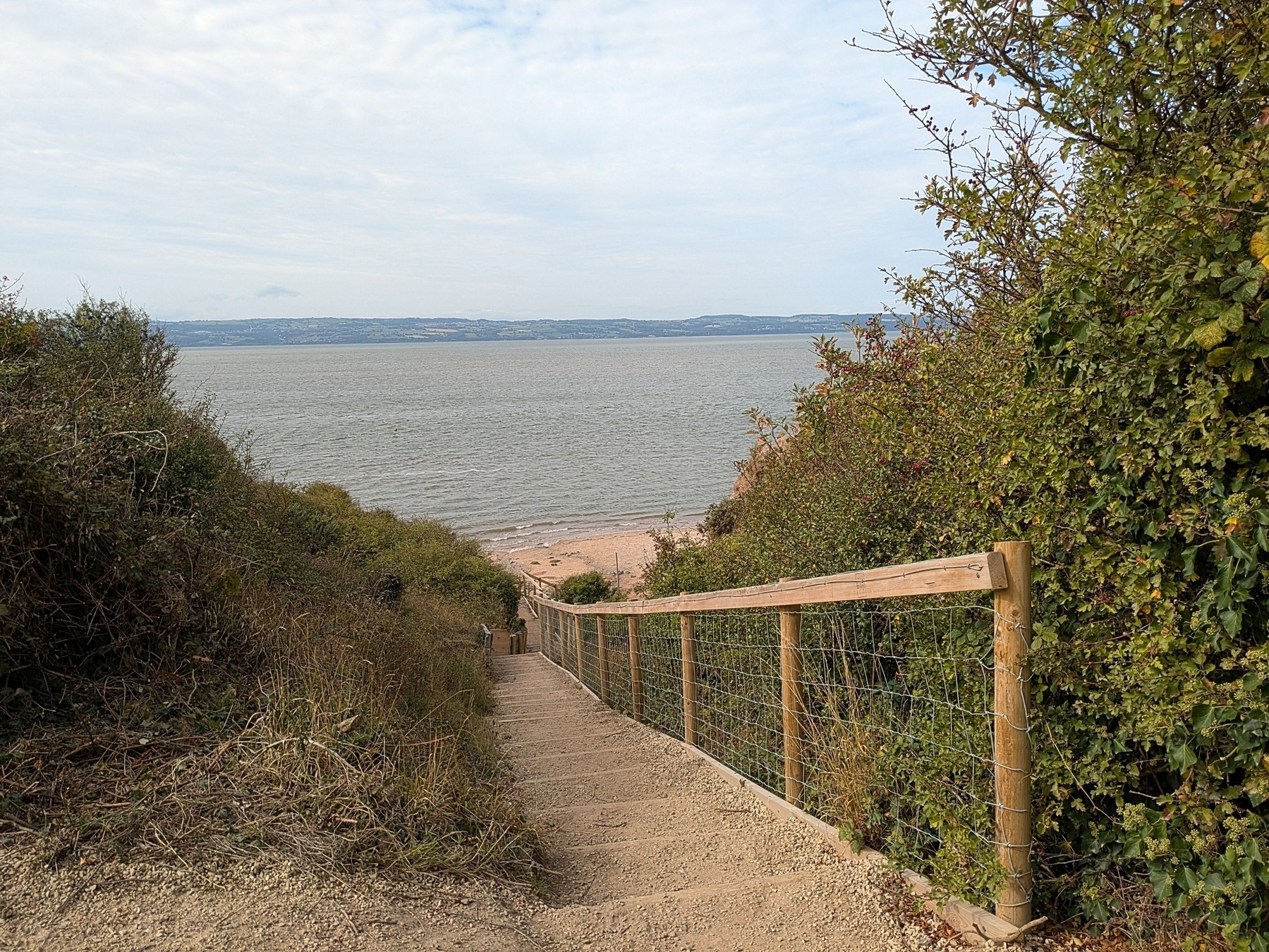 A narrow path with wooden railings leads down through greenery to a sandy beach overlooking a tranquil sea.