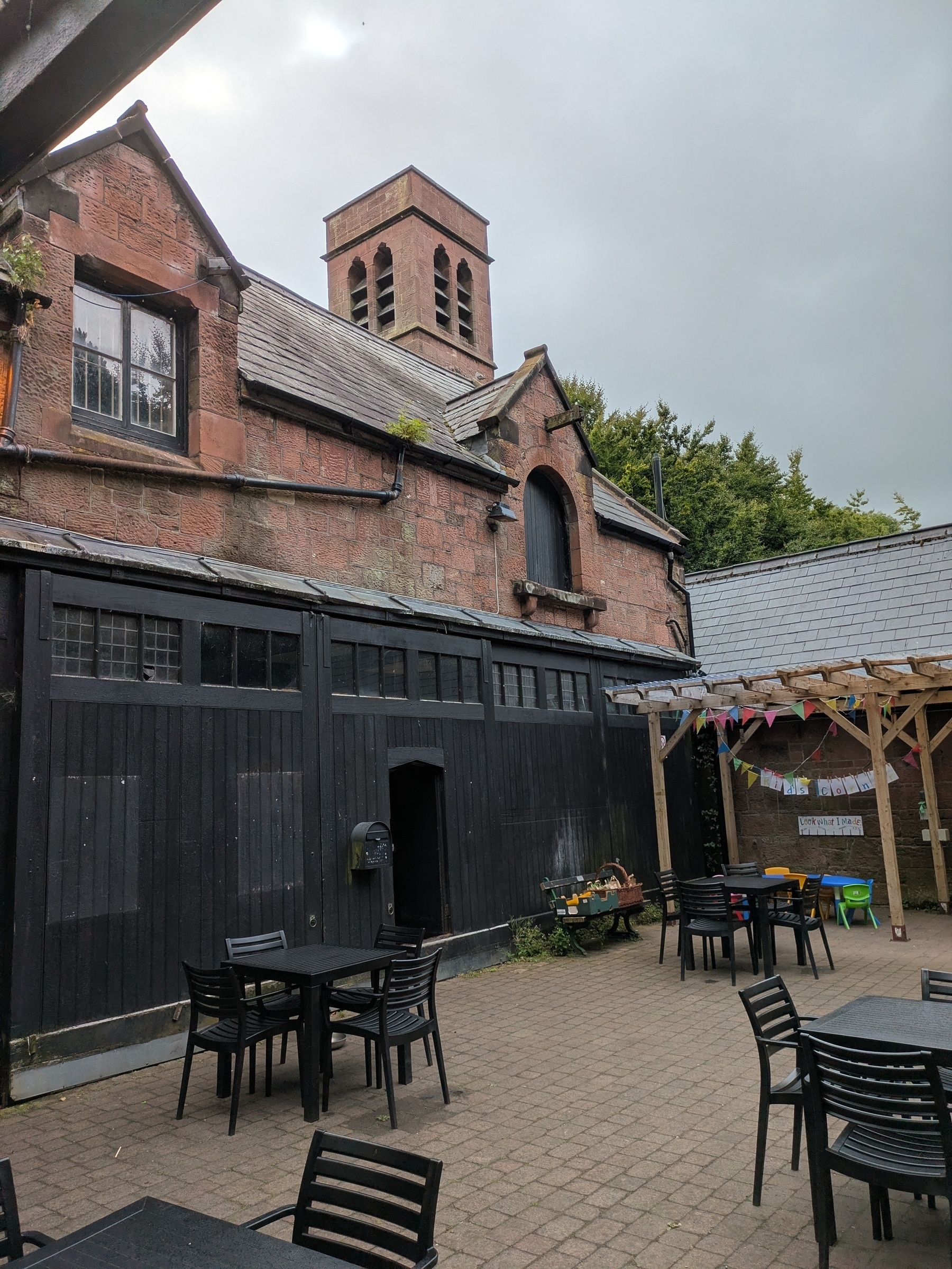A rustic outdoor seating area features black tables and chairs, surrounded by an old brick building with a small tower and a wooden gazebo adorned with colorful bunting.