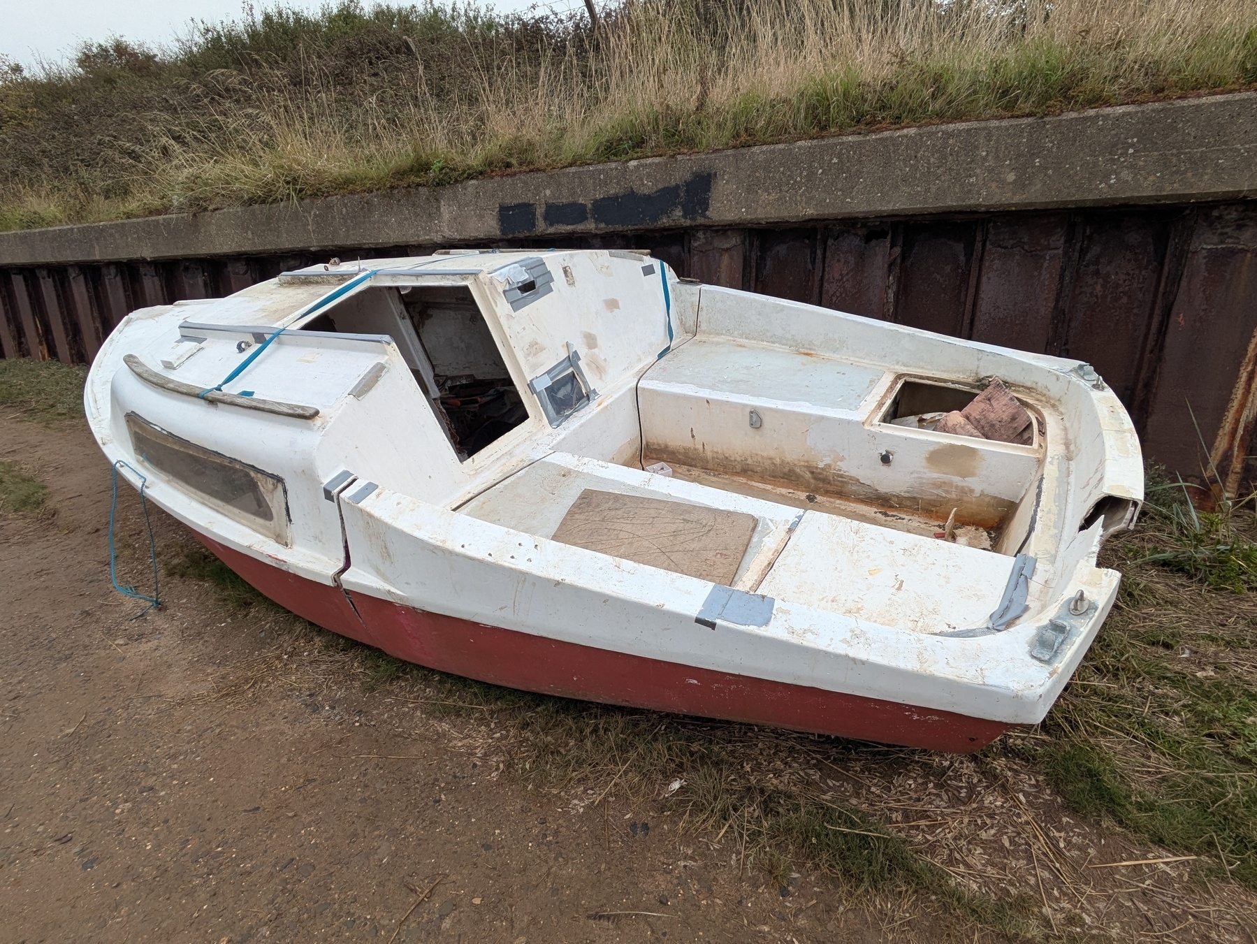 A small, weathered boat with a red hull and white deck is resting on a patch of grass near a concrete barrier.