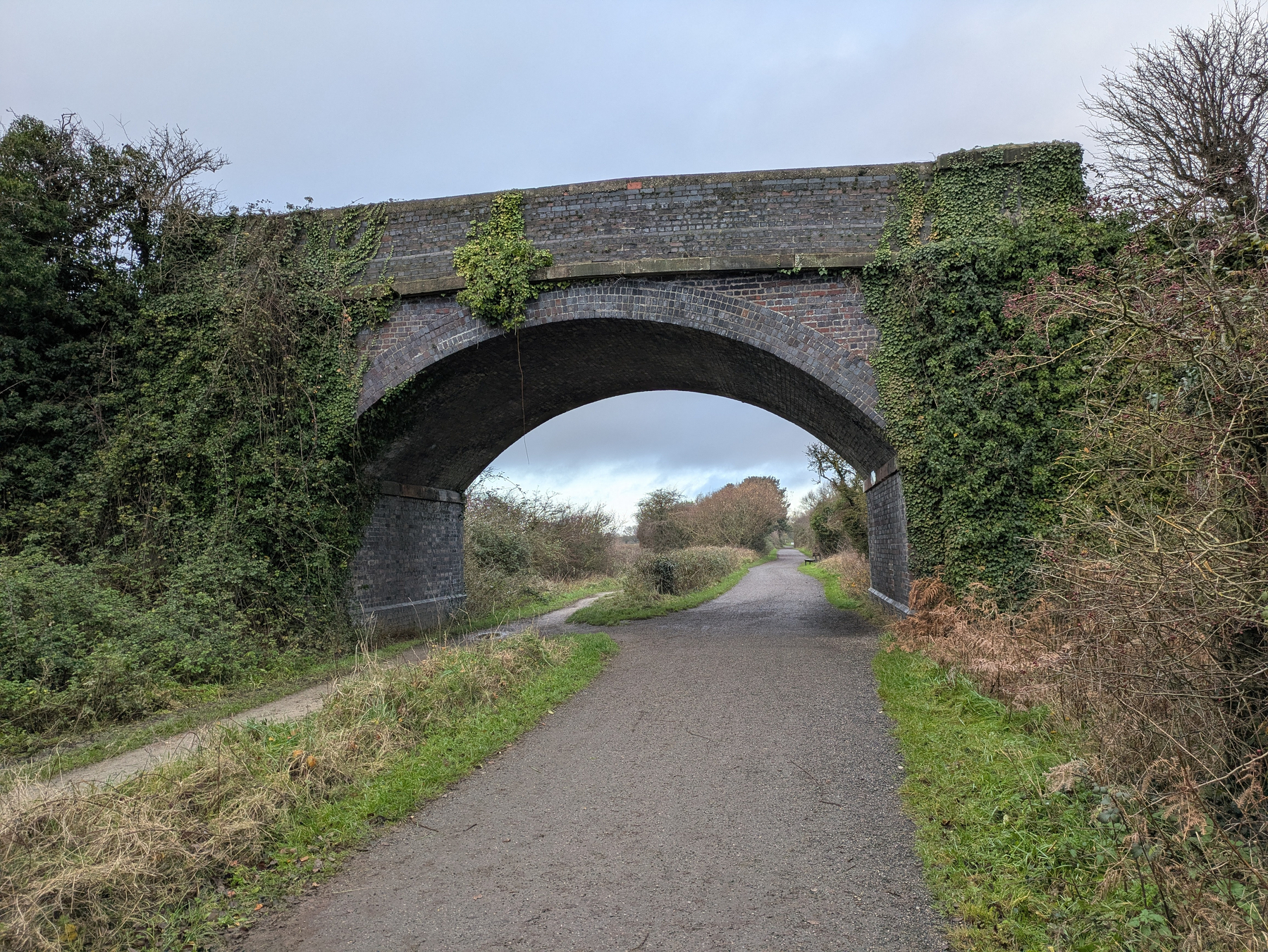 A brick bridge covered in vines spans over a pathway surrounded by greenery.