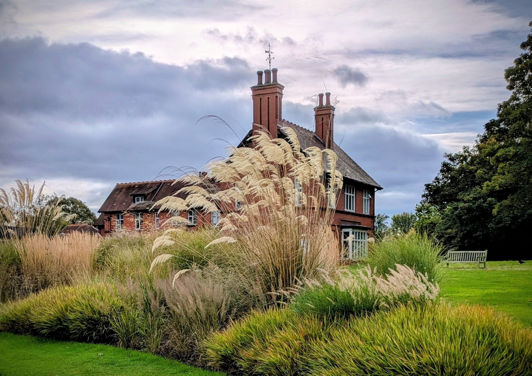 A charming brick house with tall chimneys is surrounded by lush greenery and ornamental grasses under a cloudy sky.