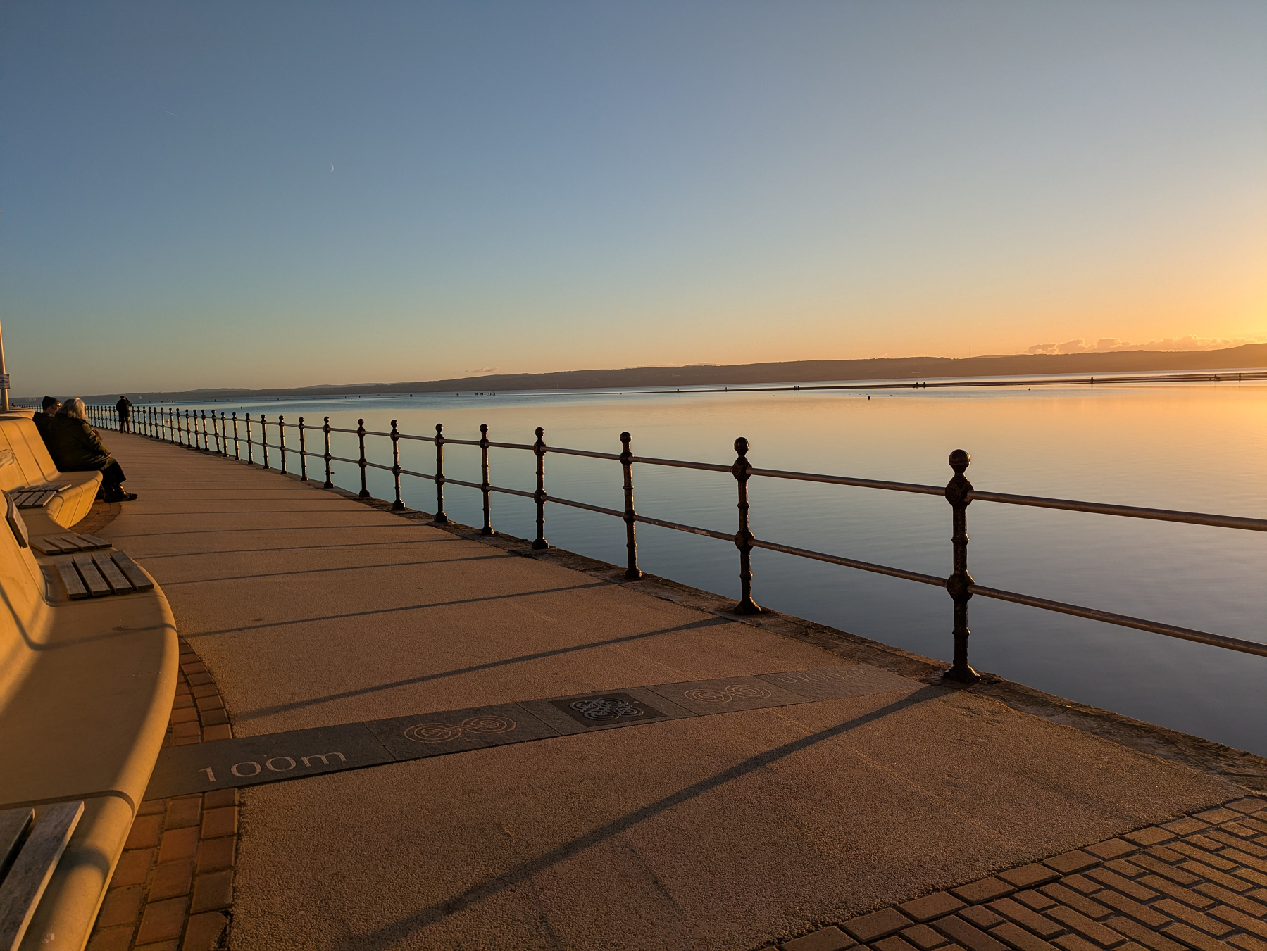 A peaceful waterfront promenade during sunset, with benches and a railing overlooking calm water.