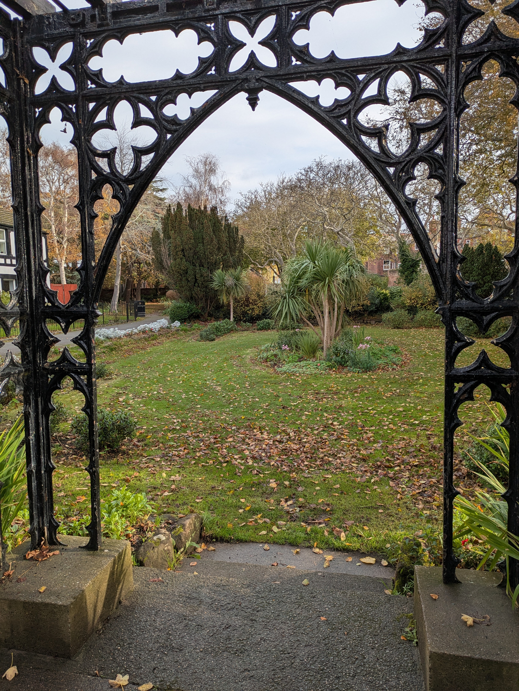 A decorative black wrought iron archway frames a leafy garden path with scattered autumn leaves and trees in the background.