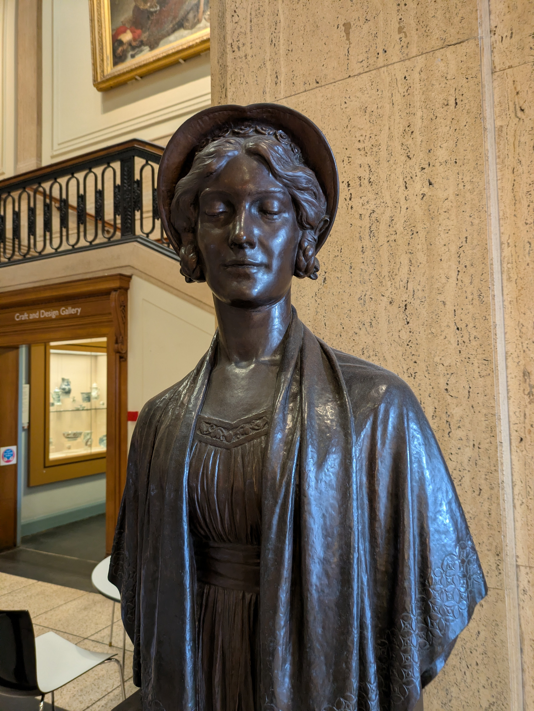 A bronze statue of a woman wearing a hat and shawl is displayed in a gallery setting with architectural details and framed artwork in the background.