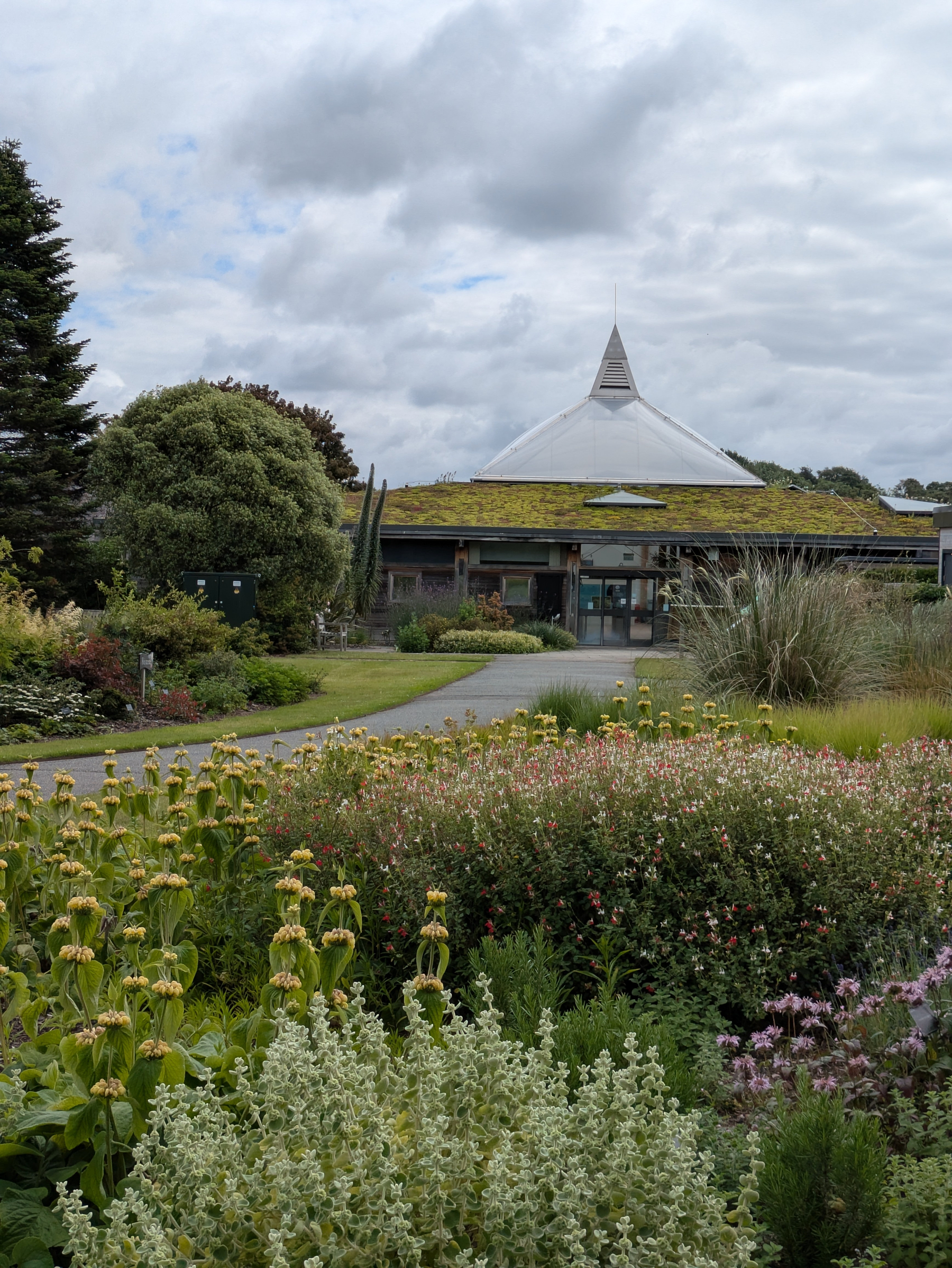 A lush garden pathway leads to a building with a unique conical roof under a cloudy sky.