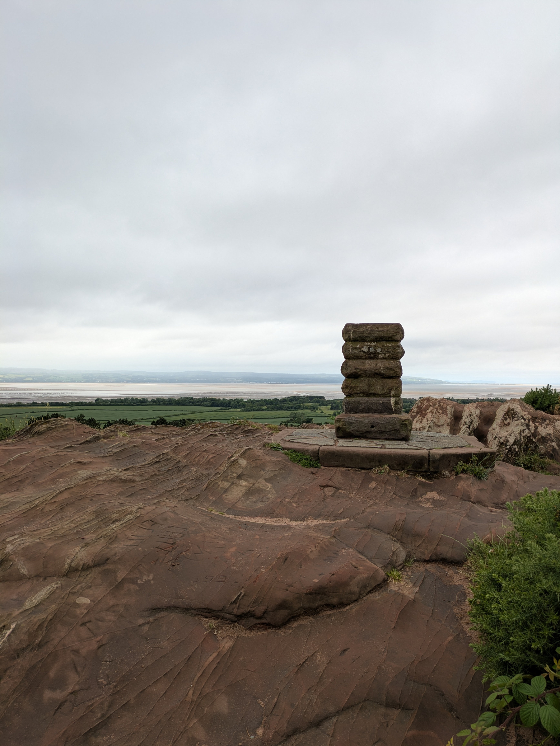 A stacked stone structure sits atop a rocky outcrop with a vast landscape and cloudy sky in the background.